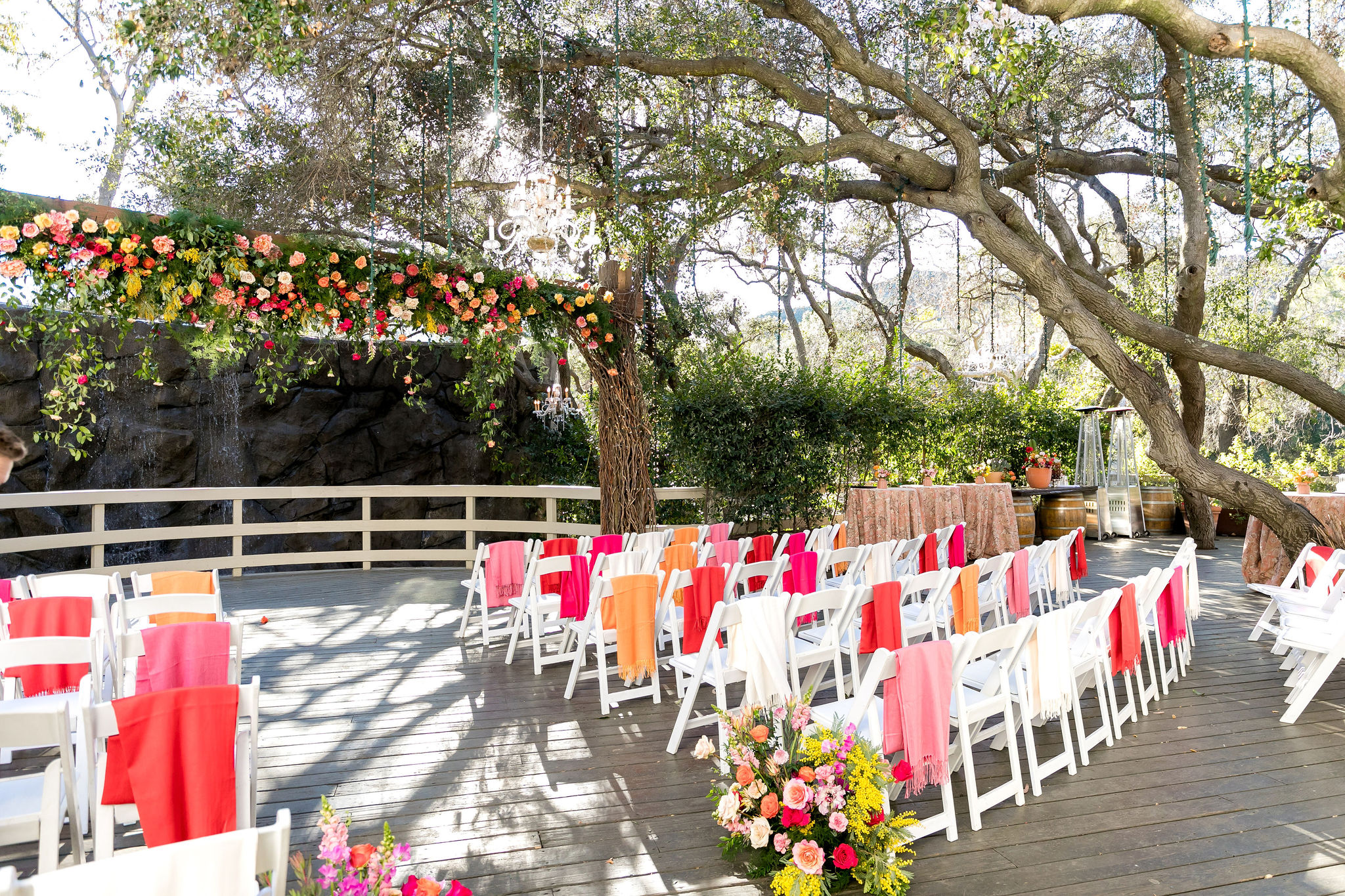 An energetic wedding ceremony at the Oak Room at Calamigos Ranch ceremony space with bright orange, red, pink and white florals and shawls hanging from ceremony chairs