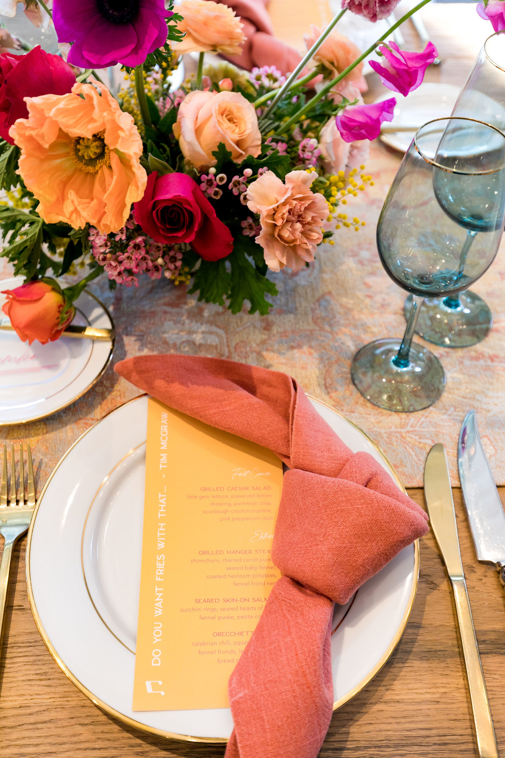 bright and energetic wedding reception tables with orange napkins and menu, with red, purple, orange and pink floral centerpieces on wooden farm tables