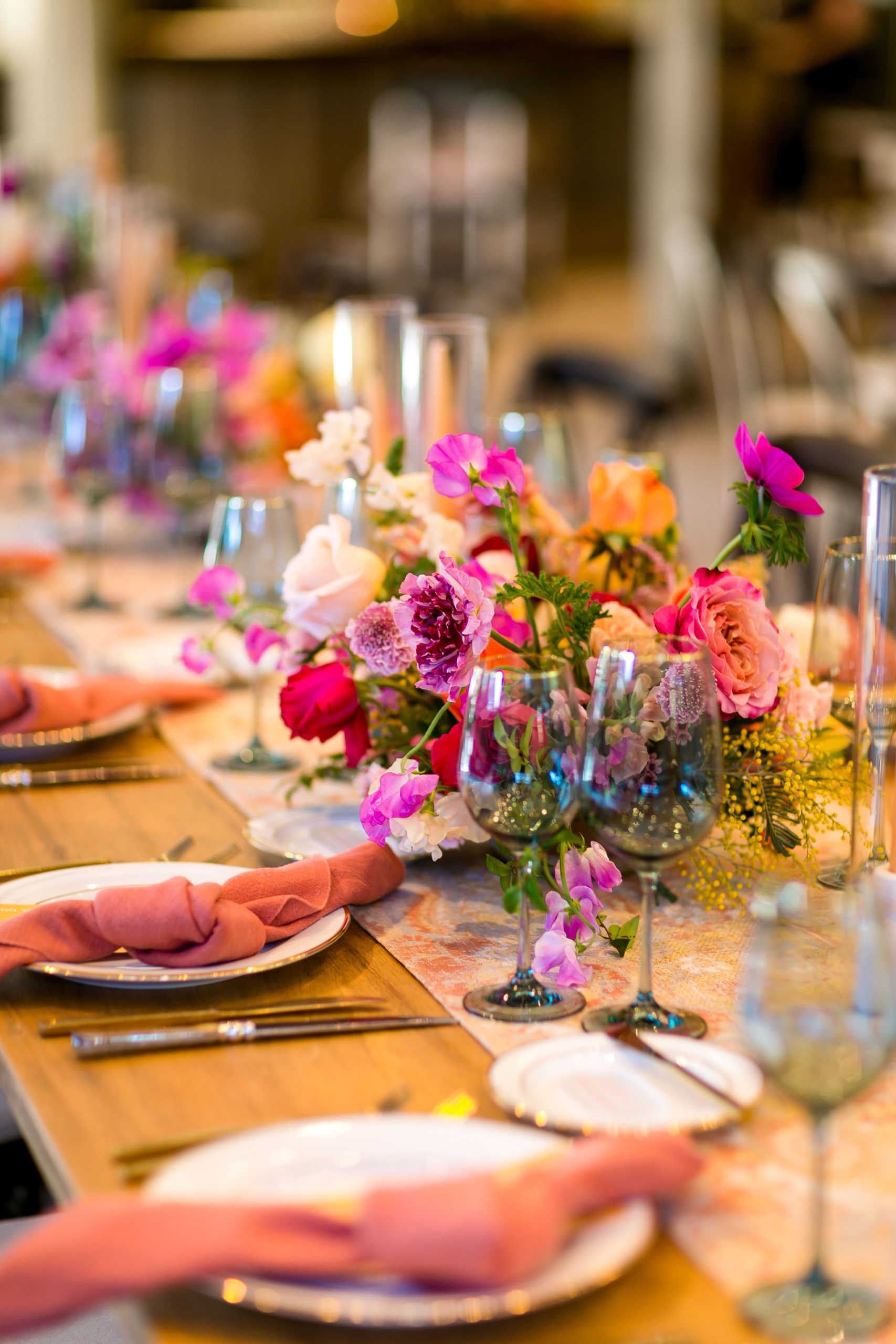 bright and energetic wedding reception tables with orange napkins and red, purple, orange and pink floral centerpieces on wooden farm tables