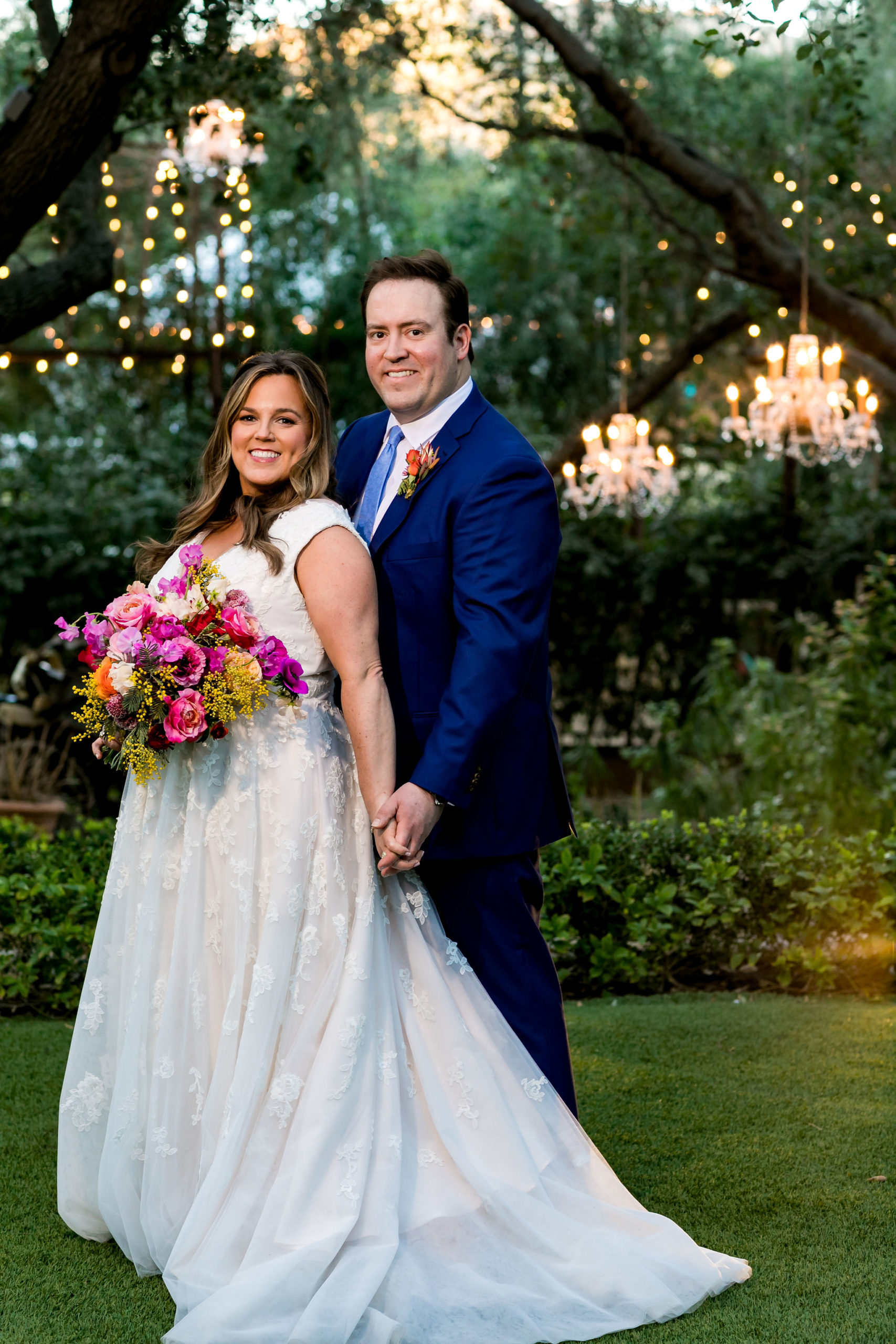 bride in v-neck wedding dress with lace overlay holding a bright purple and pink bouquet stands with groom in bright blue suit and tie