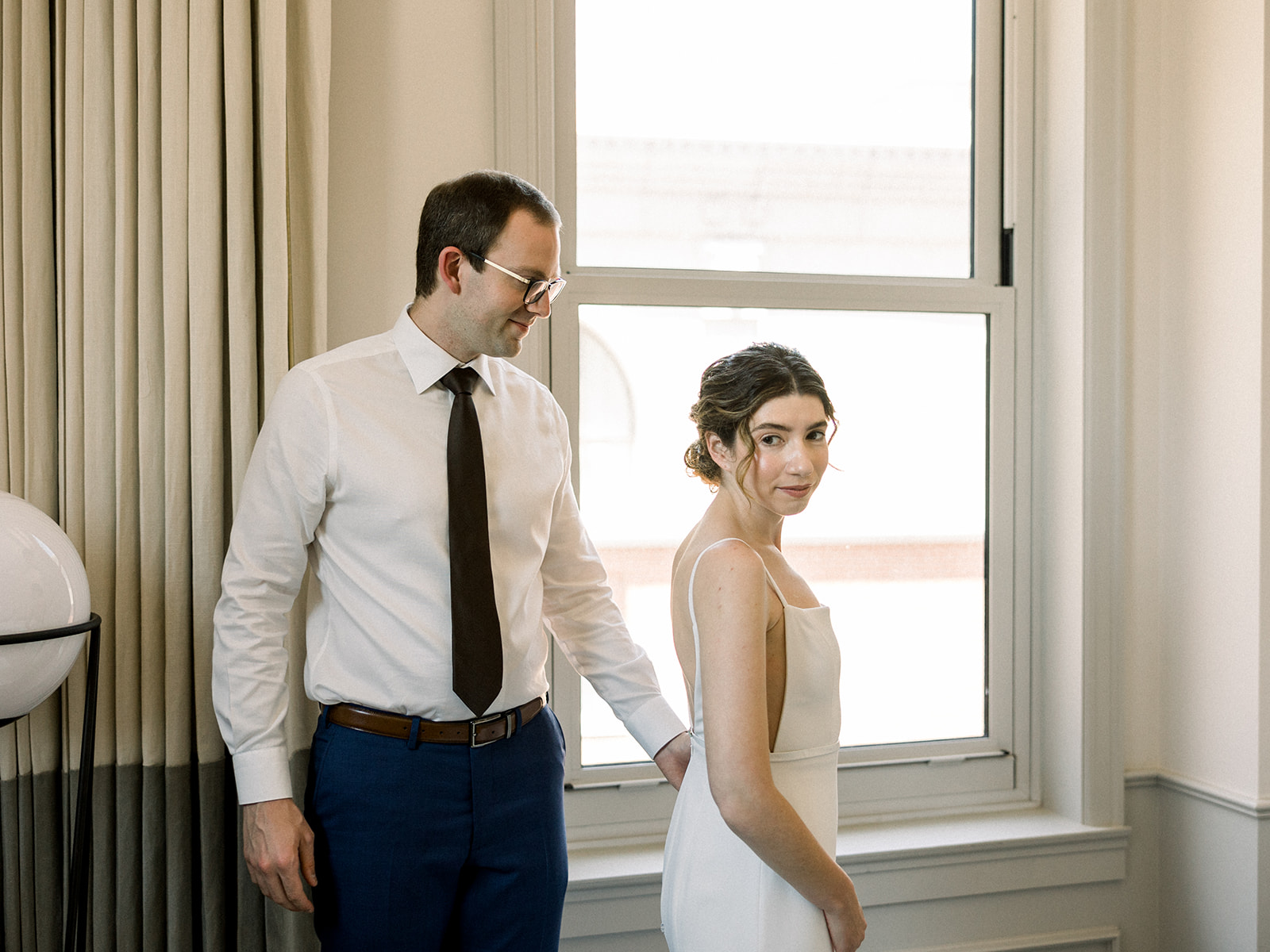 bride in contemporary spaghetti strap dress stands with groom in white shirt and navy pants