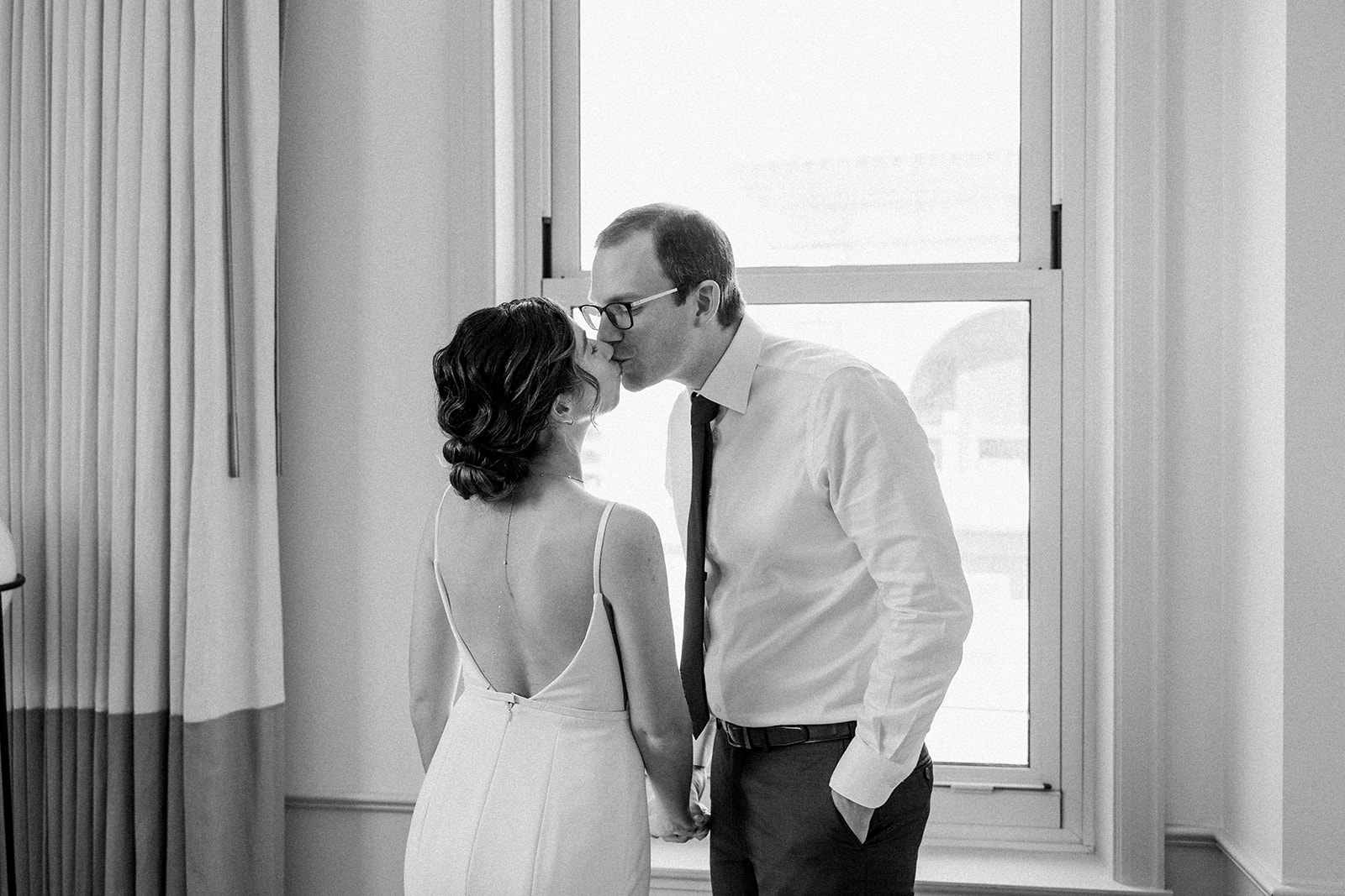 bride in contemporary spaghetti strap dress and low chignon bun kisses groom wearing glasses