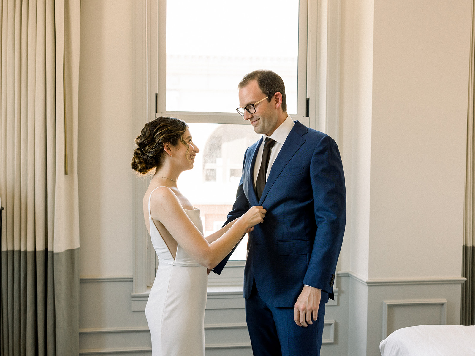bride in contemporary spaghetti strap dress and low chignon bun poses with groom wearing glasses and navy suit