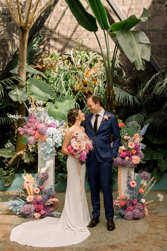 bride in contemporary wedding dress stands with groom in dark blue suit in front of ceremony arch made of pastel florals and greenery at the Grass Room in DTLA