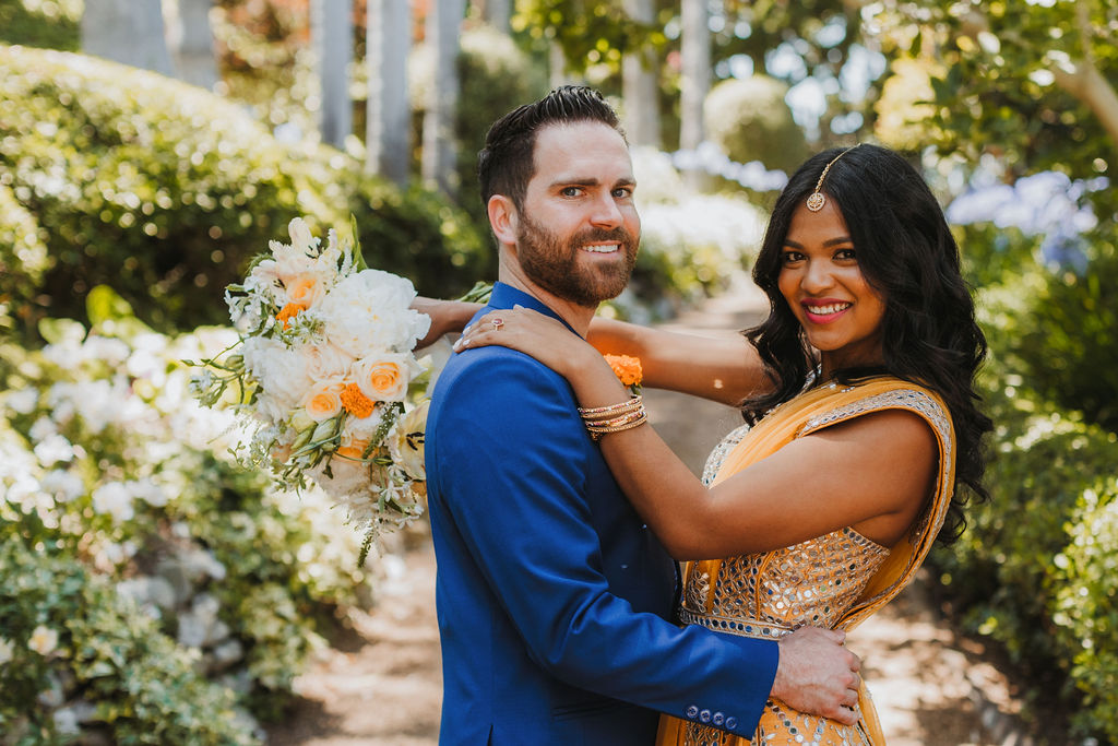 bride in orange marigold colored wedding sari holds bouquet of white and orange flowers during portraits with groom in cobalt blue suit and orange marigold boutonniere at Lake Shrine Temple