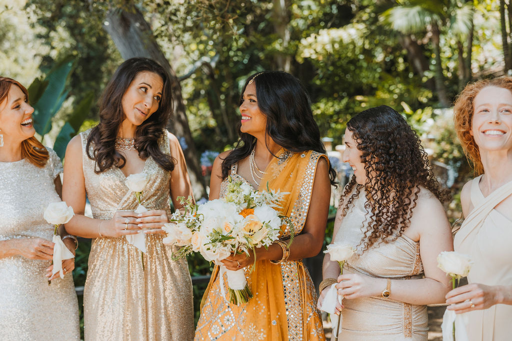 bride in orange marigold colored wedding sari holds bouquet of white and orange flowers during portraits with bridesmaids in mix matched gold and beige bridesmaid dresses