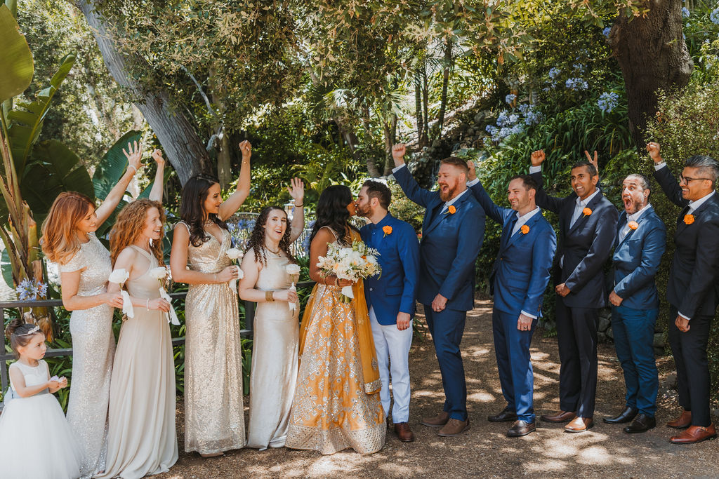 bride in orange marigold colored wedding sari holds bouquet of white and orange flowers with groom in cobalt blue suit stands with wedding party wearing mix matched gold and beige bridesmaid dresses and blue and grey suits at Lake Shrine Temple