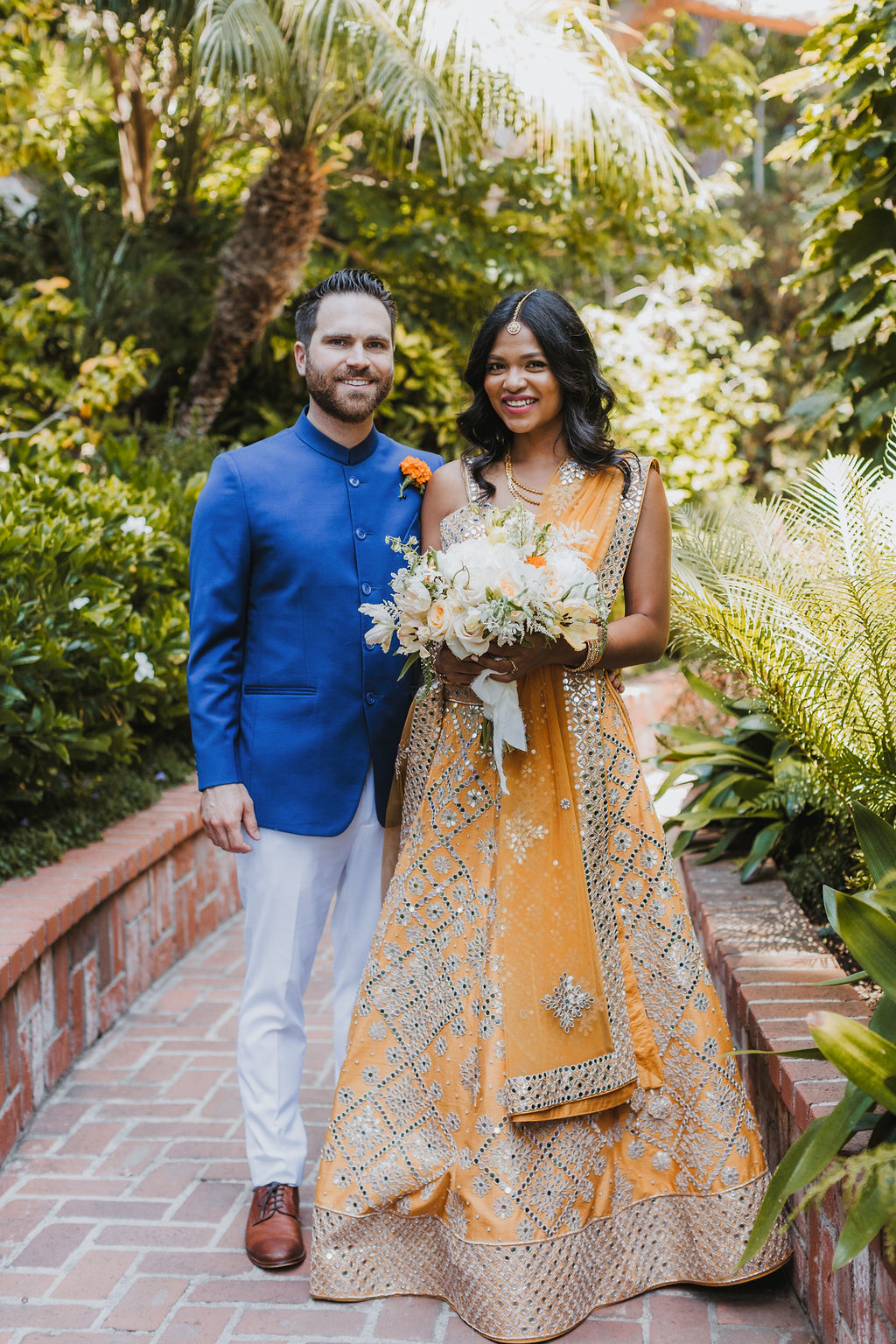 bride in orange marigold colored wedding sari holds bouquet of white and orange flowers with groom in cobalt blue suit before their wedding at Lake Shrine Temple