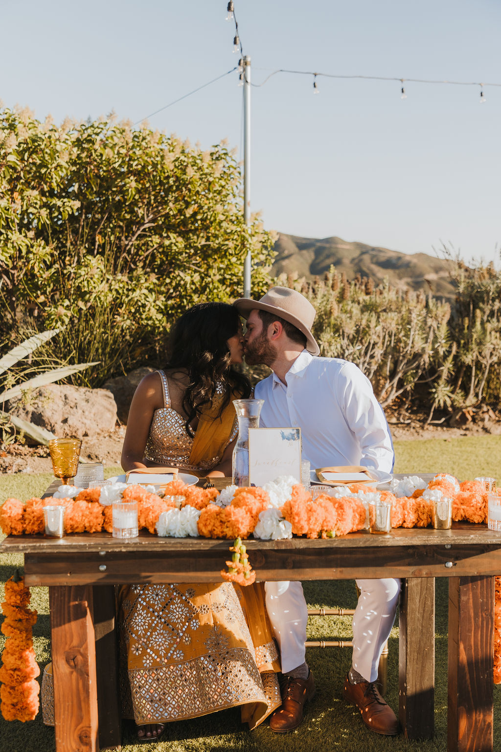 bride in orange sari and groom in white shirt and hat kiss at sweetheart table during wedding reception at Saddlerock Ranch in Malibu