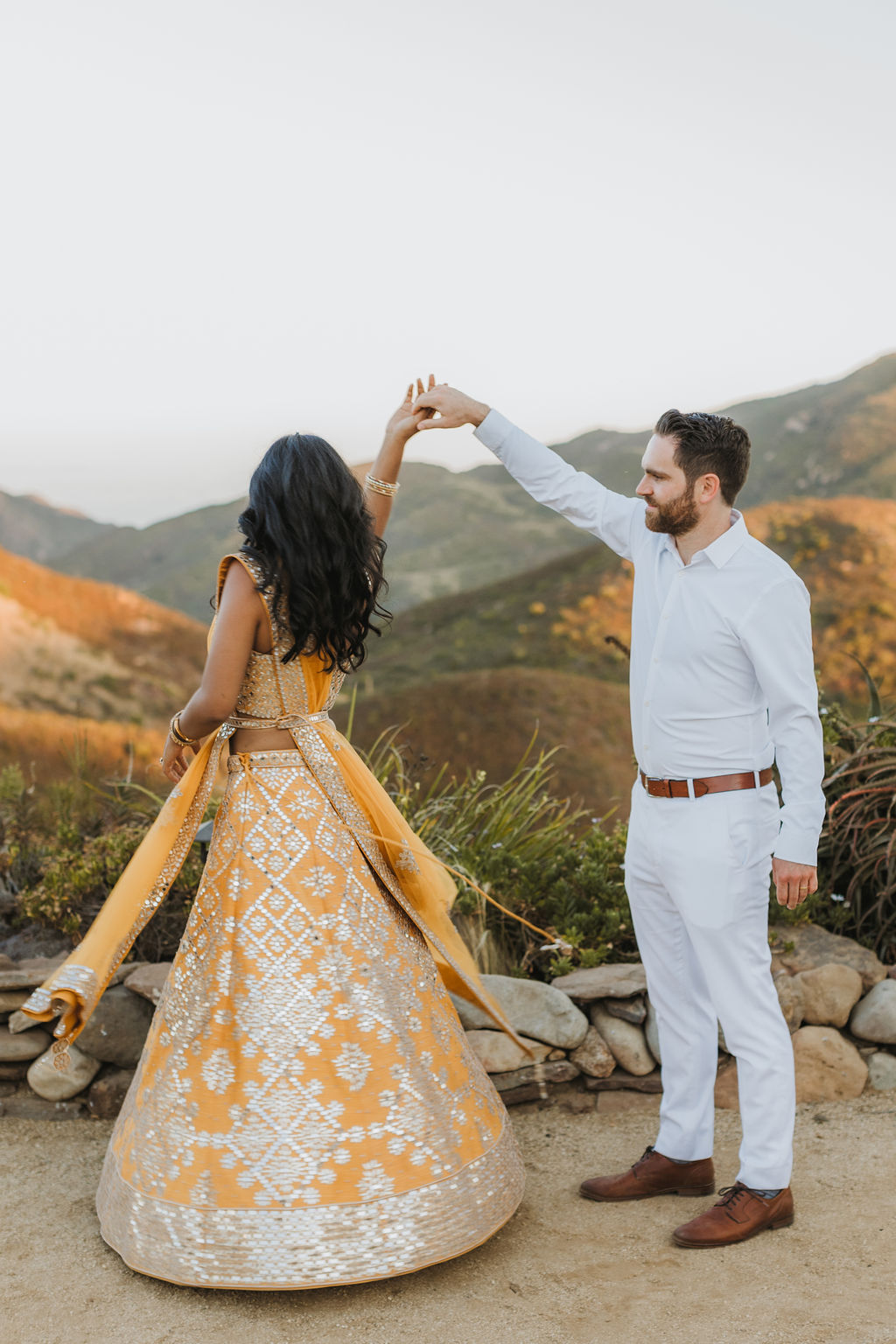 bride in orange sari dances with groom in white during sunset photos at Saddlerock Ranch in Malibu