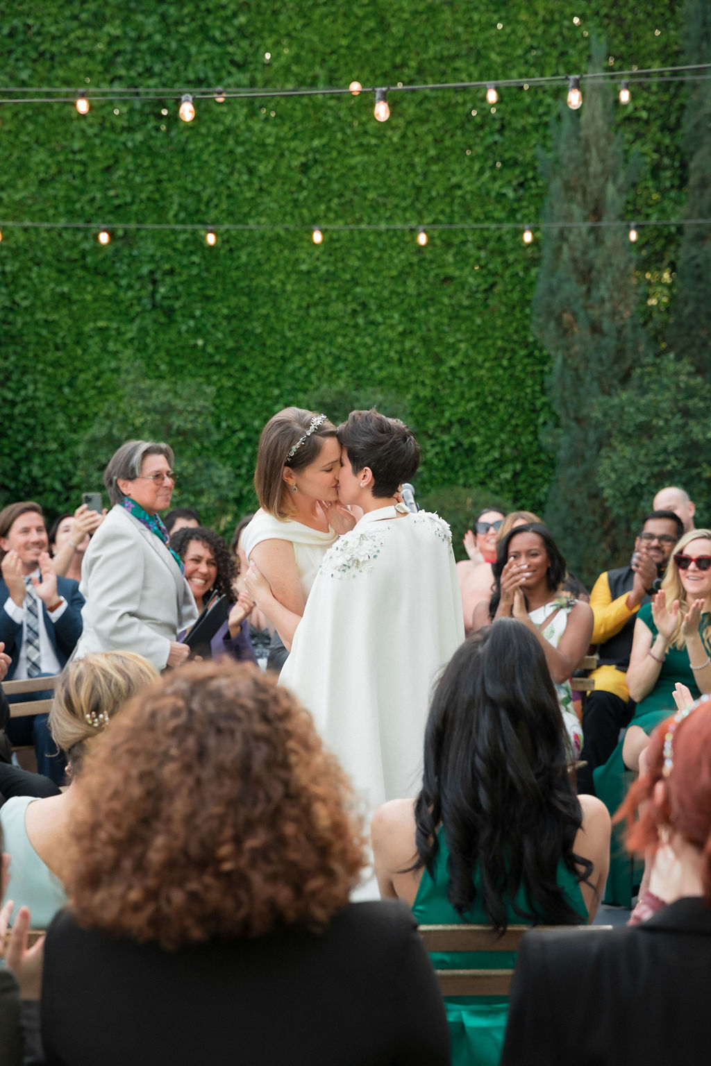 two brides wearing coordinating bridal attire share first kiss during outdoor wedding ceremony at the Fig House