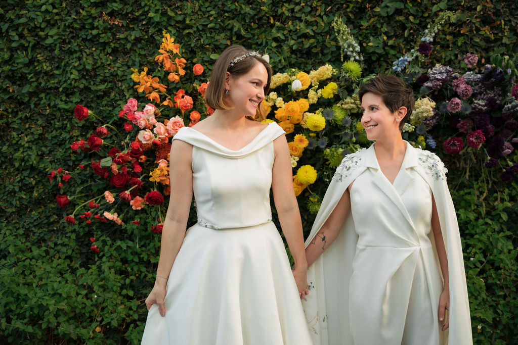 brides in coordinating bridal dress and bridal jumpsuit hold hands in front of hanging rainbow floral installation after wedding ceremony at The Fig House