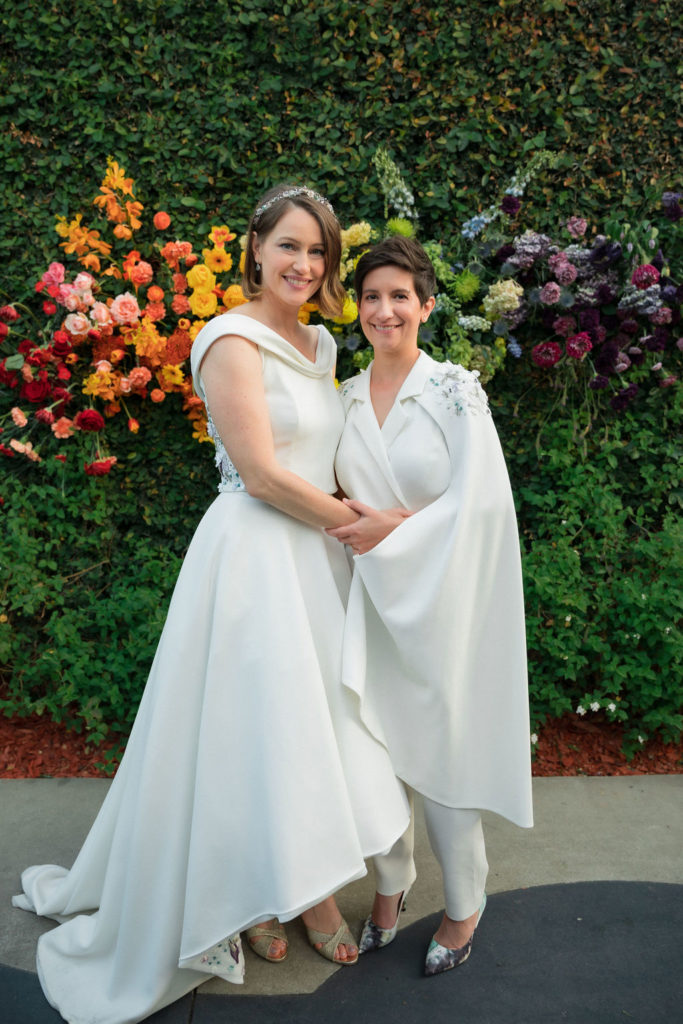 Brides in custom made coordinated wedding outfits stand in front of rainbow hanging floral arrangement after ceremony at Fig House