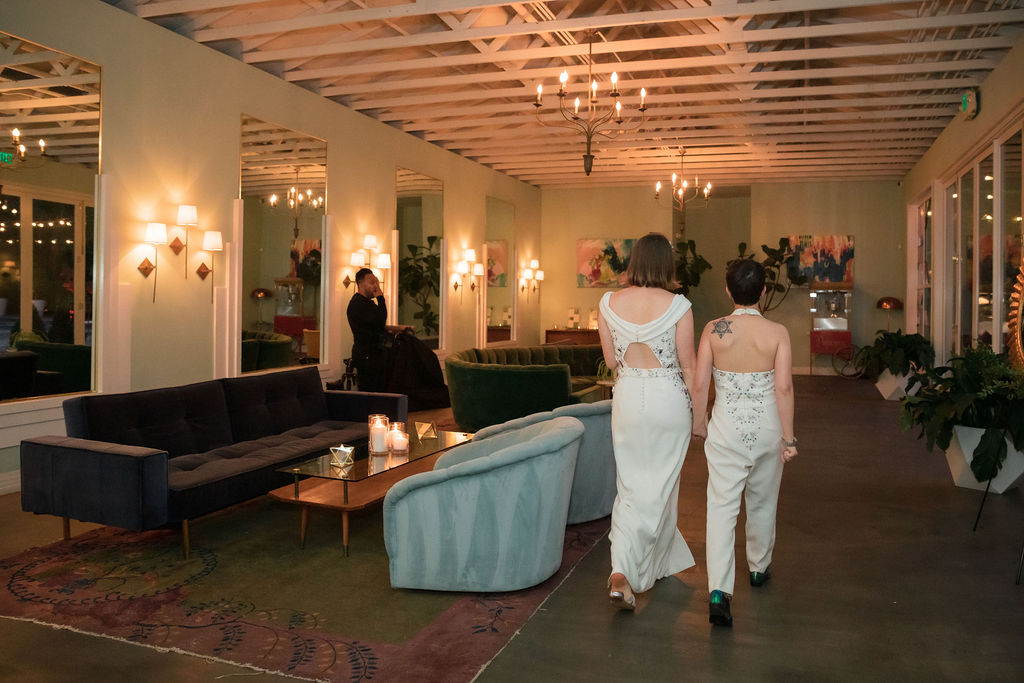 two brides in coordinated wedding dress and bridal jumpsuit hold hands as they leave their wedding reception 