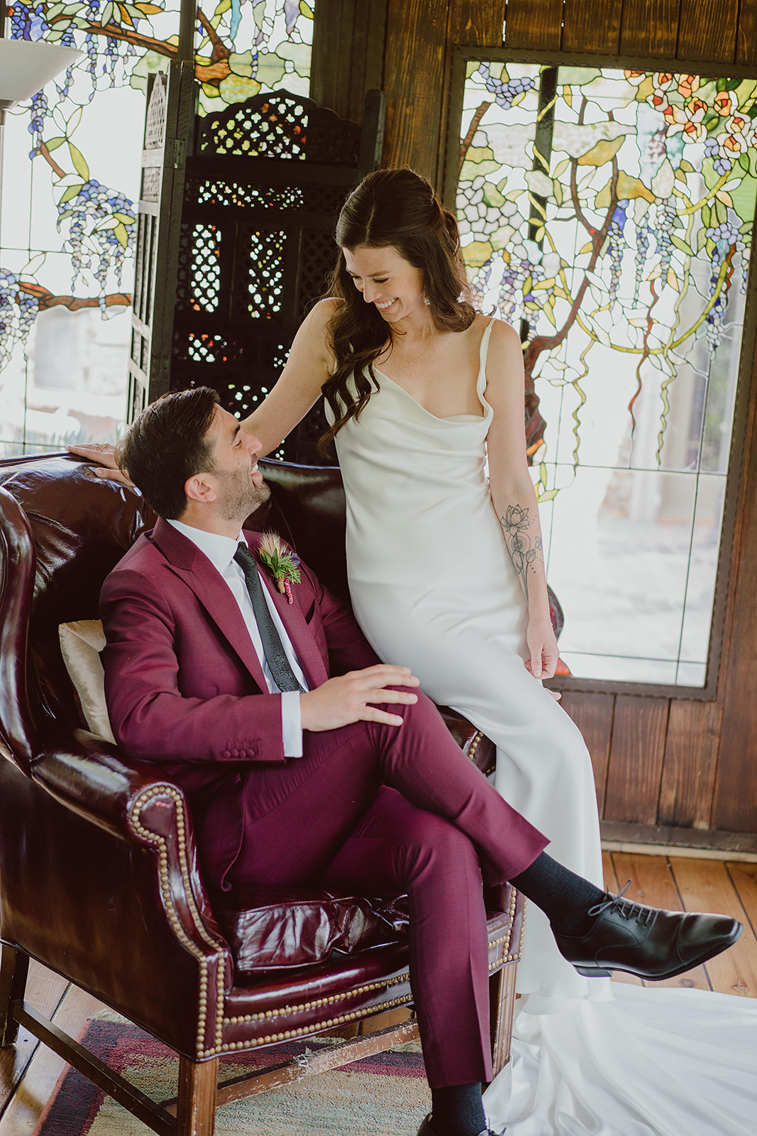 bride in minimalist silk wedding dress sits with groom in burgundy suit in a leather chair before their magical wedding at The Houdini Estate in Hollywood