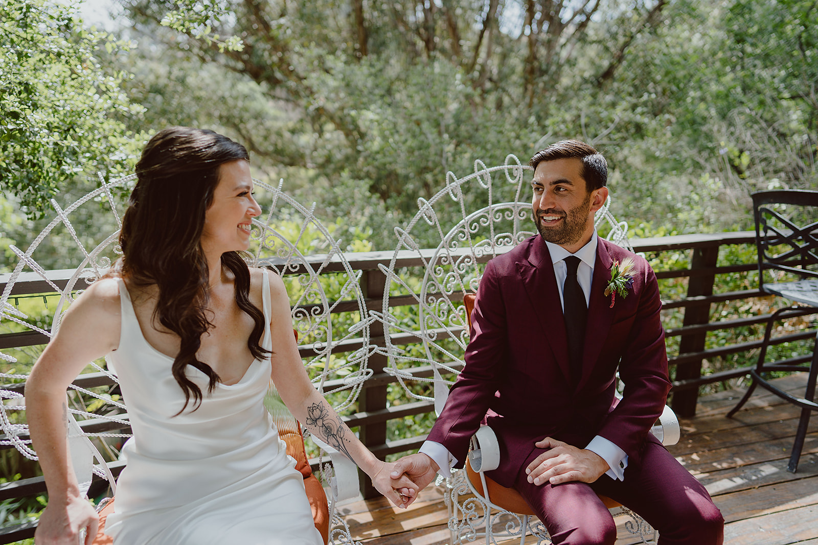 bride in minimalist satin wedding dress sits in chair with groom in burgundy suit and black tie before their magical wedding at The Houdini Estate in Hollywood