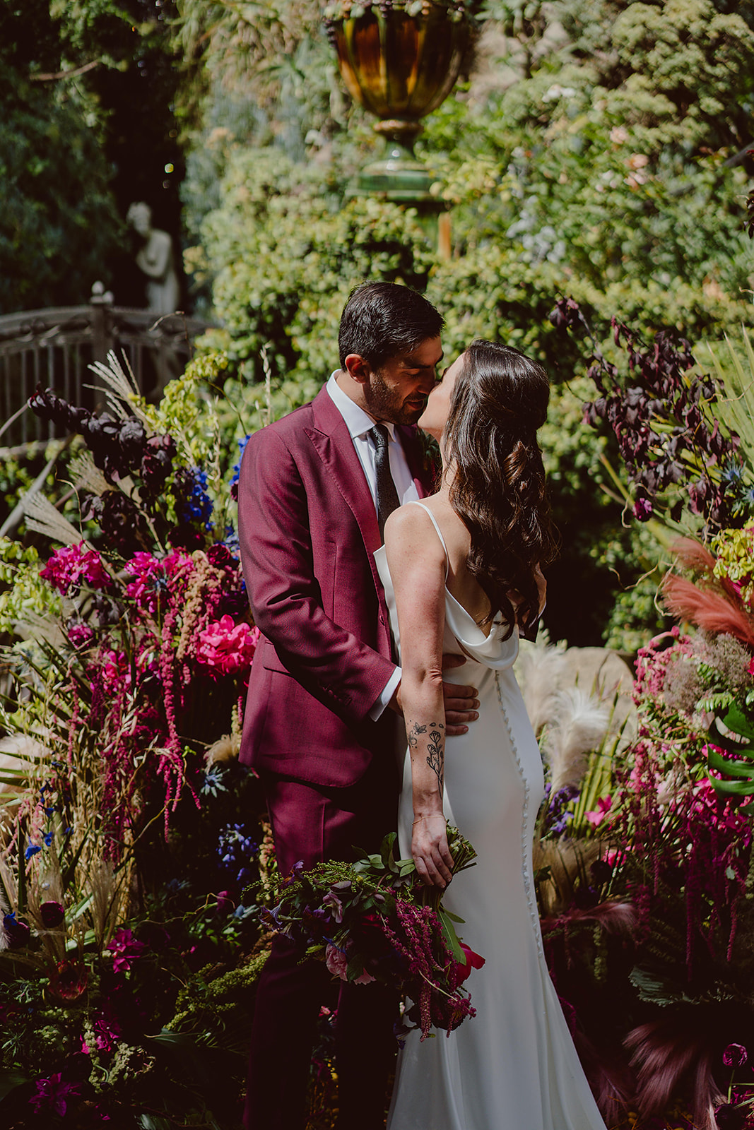 minimalist bride in satin wedding dress kisses groom wearing dark burgundy suit 