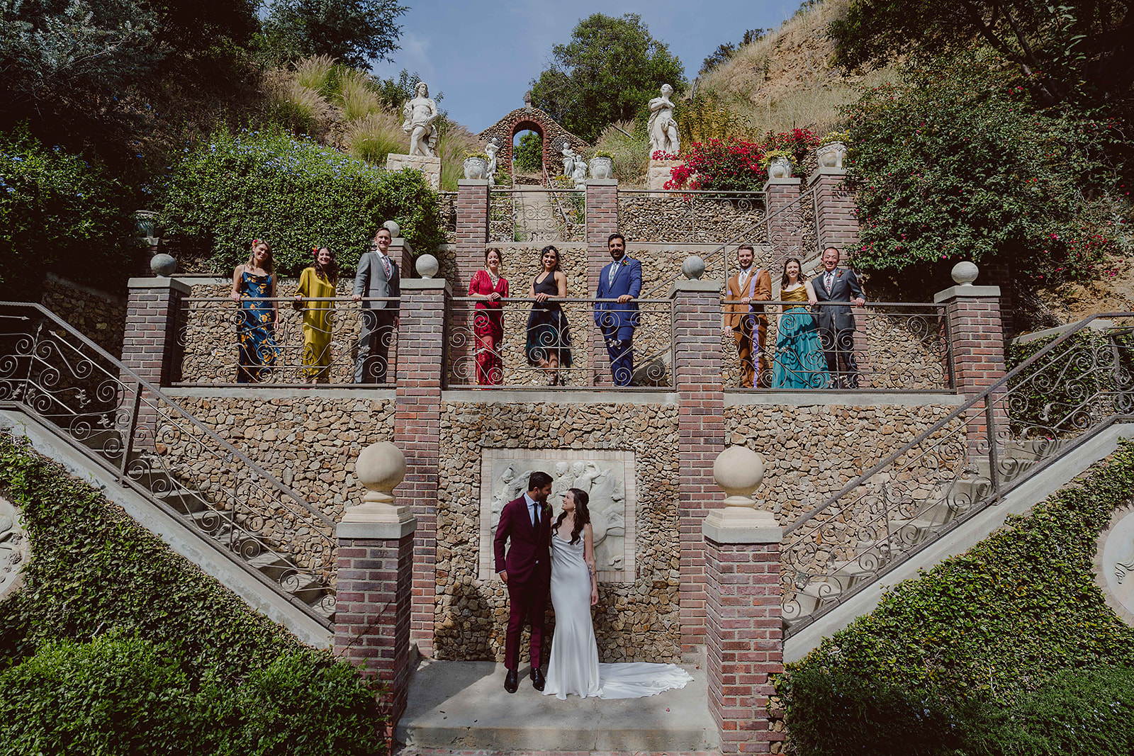  minimalist bride in satin wedding dress stands with groom in Burgundy suit at bottom of staircase with wedding party in rainbow apparel at the top of the stairs