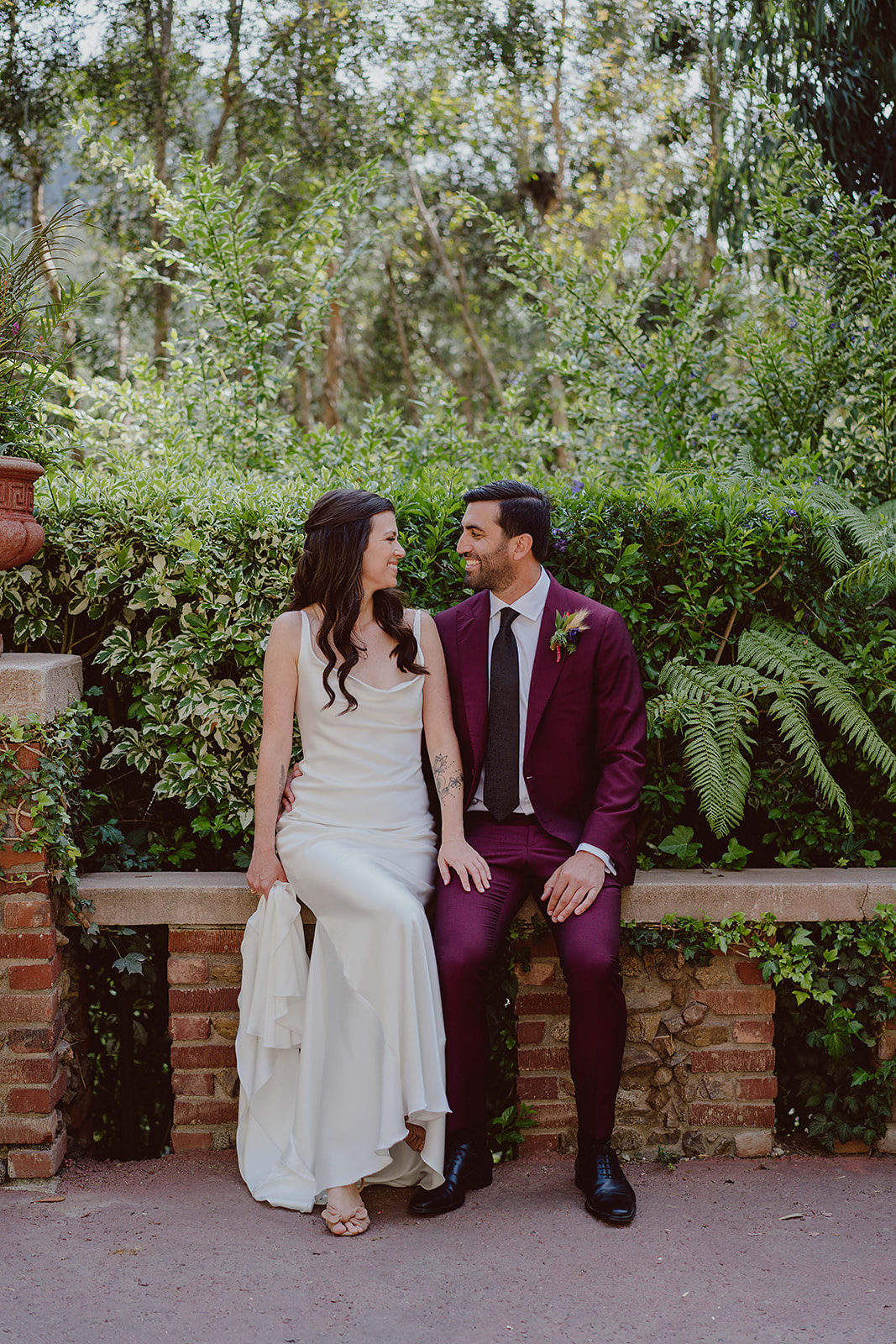 minimalist bride in silk dress sits on stone bench with groom in burgundy suit and black tie before their magical wedding at The Houdini Estate in Hollywood