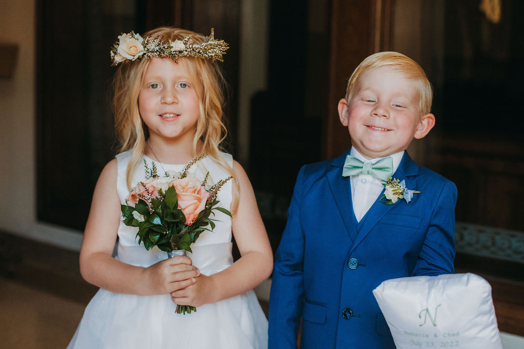 flower girl with small bouquet and floral crown stands next to ring bearer with blue suit and green bowtie 