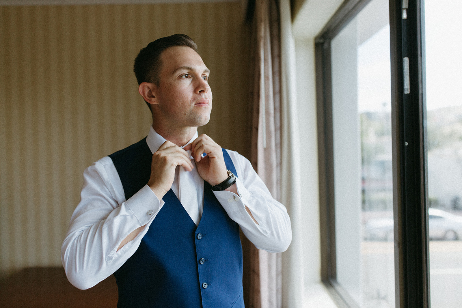 groom in blue vest and white shirt with white tie gets ready for wedding day