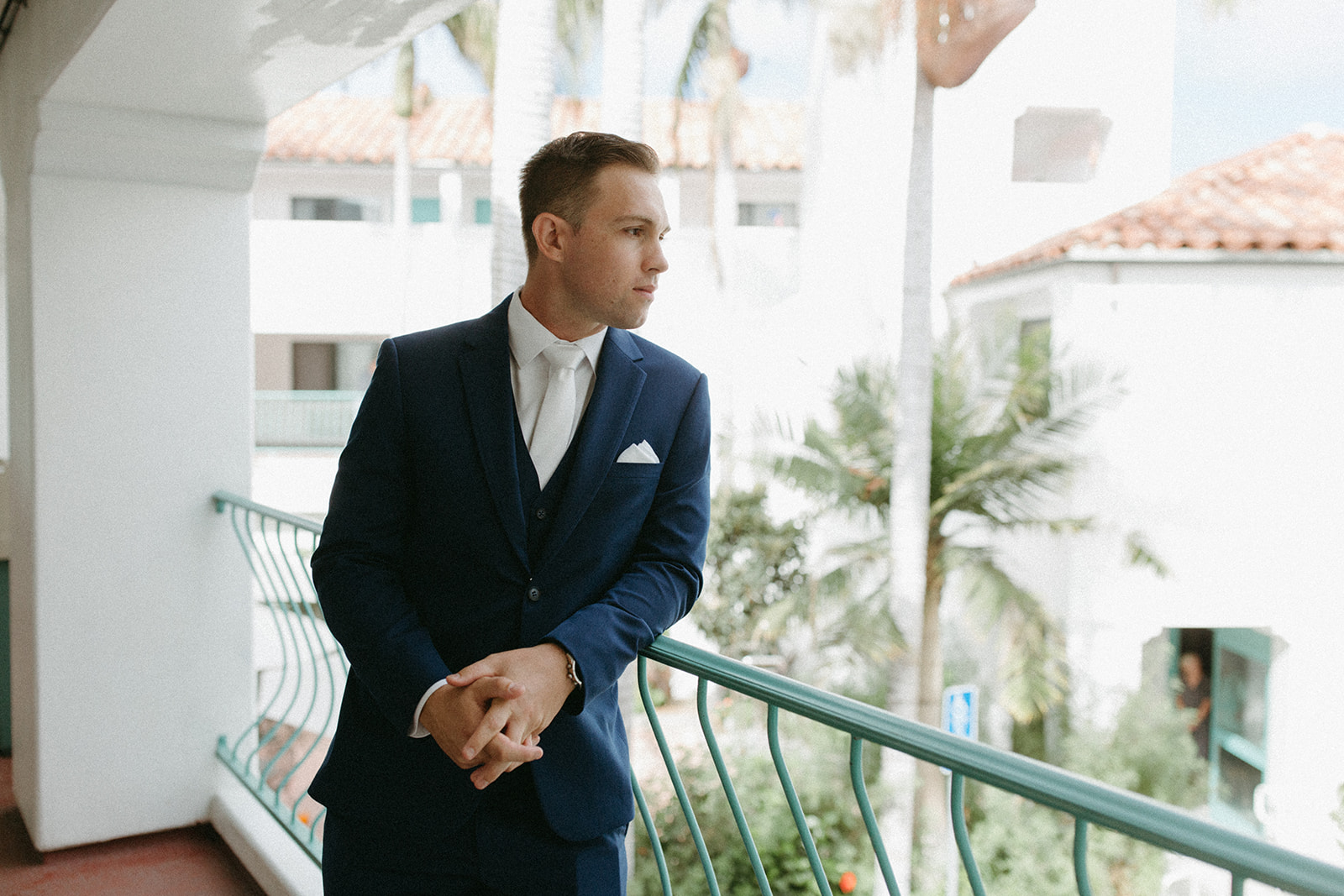 groom in blue suit with white tie stands on balcony 