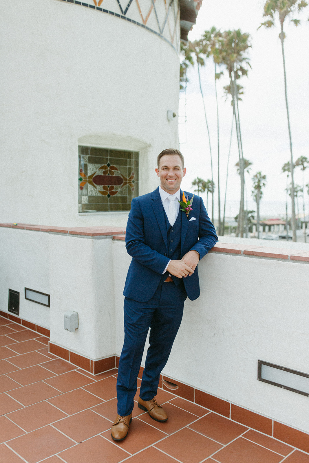 groom in blue suit and white tie with brown shoes and a colorful tropical boutonniere 