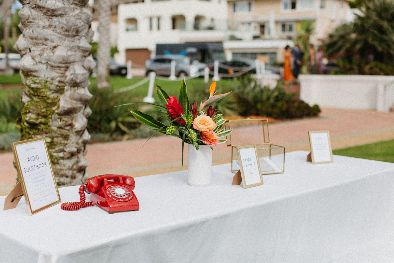 wedding welcome table with tropical flower arrangement, red vintage telephone as an audio guest book and gold framed glass card box