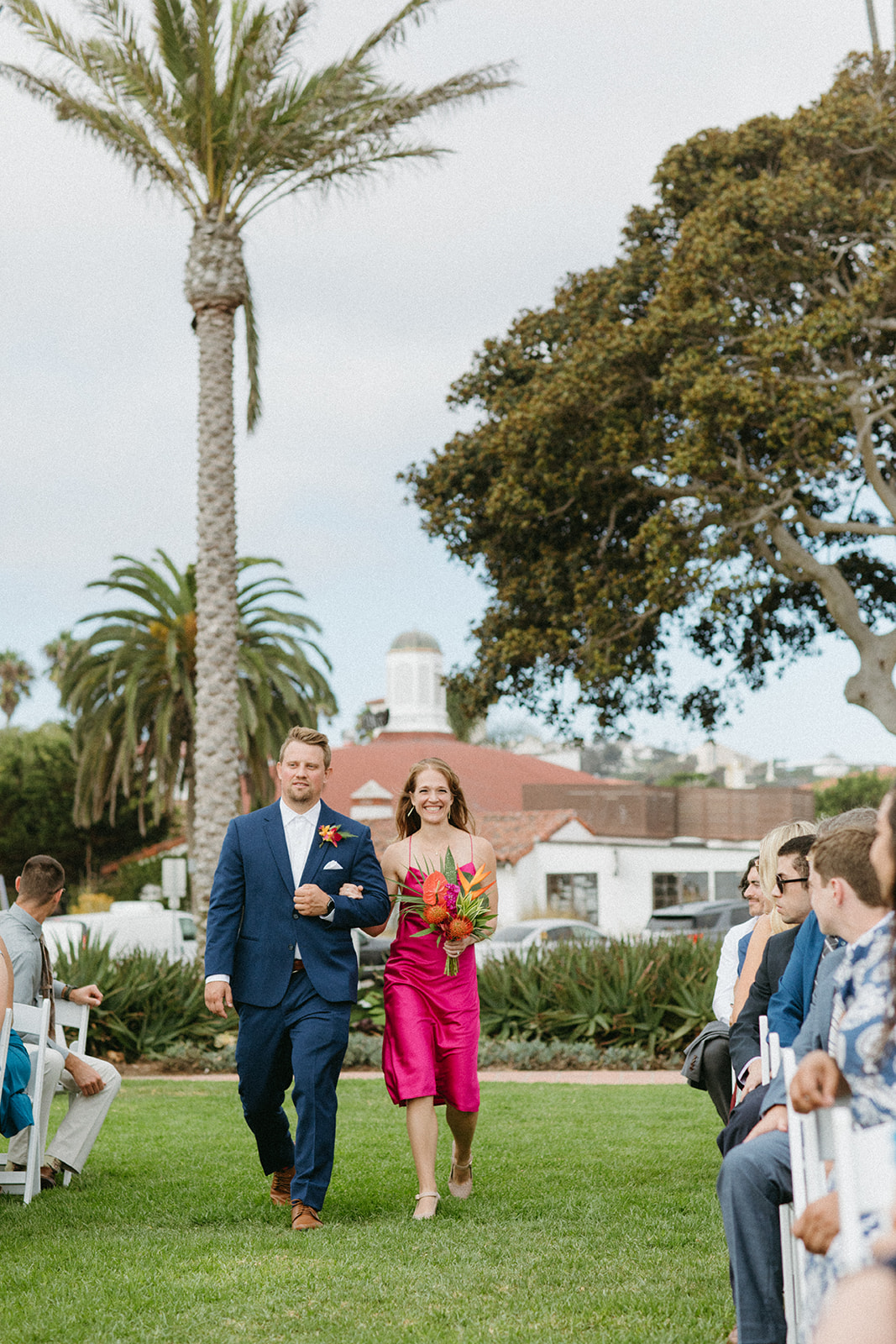 wedding party with groomsman in blue suit and bridesmaid in bright pink dress with tropical bouquet walk down aisle
