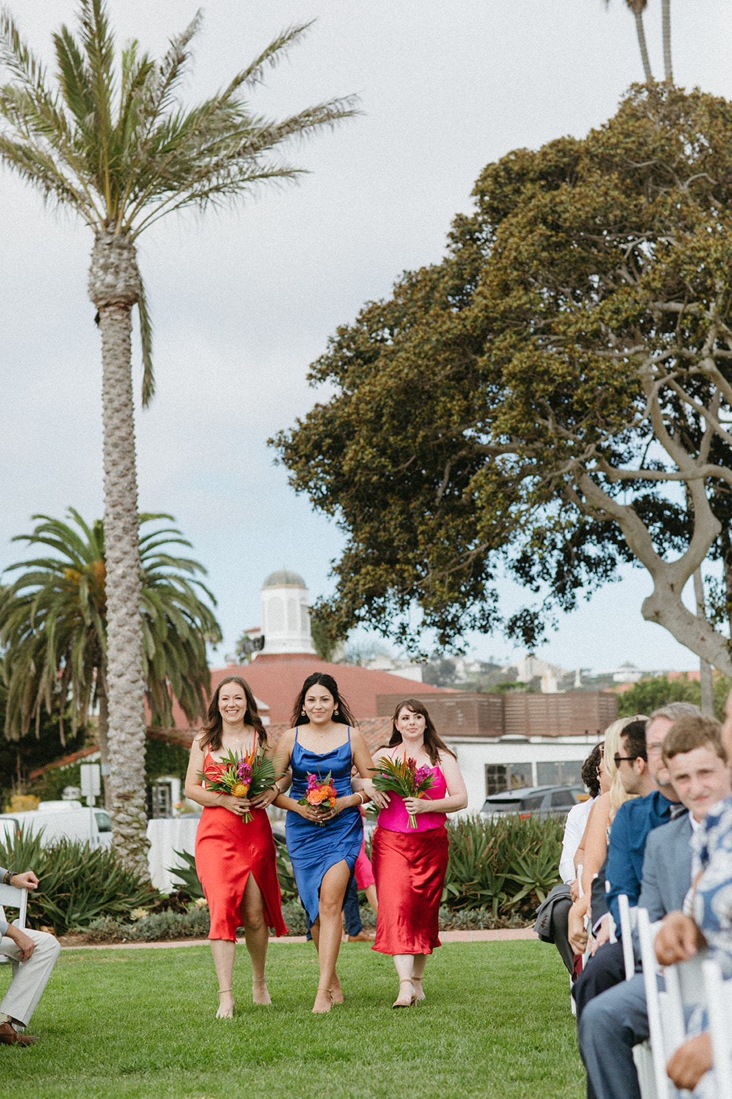 bridesmaids in bright pink, blue and red dresses with tropical bouquet walk down aisle