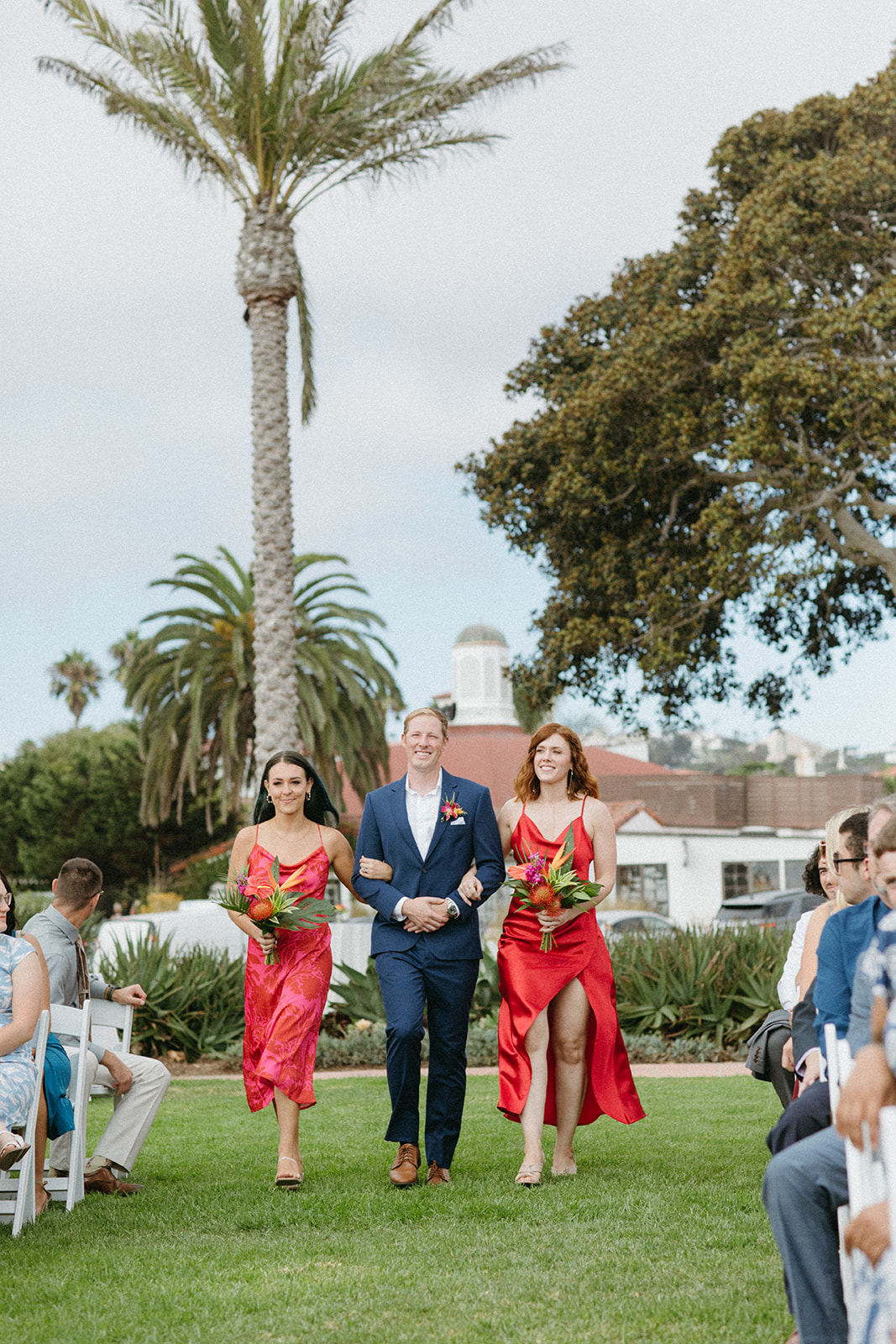 wedding party with groomsman in blue suit and bridesmaids in bright pink dresses with tropical bouquets walk down aisle