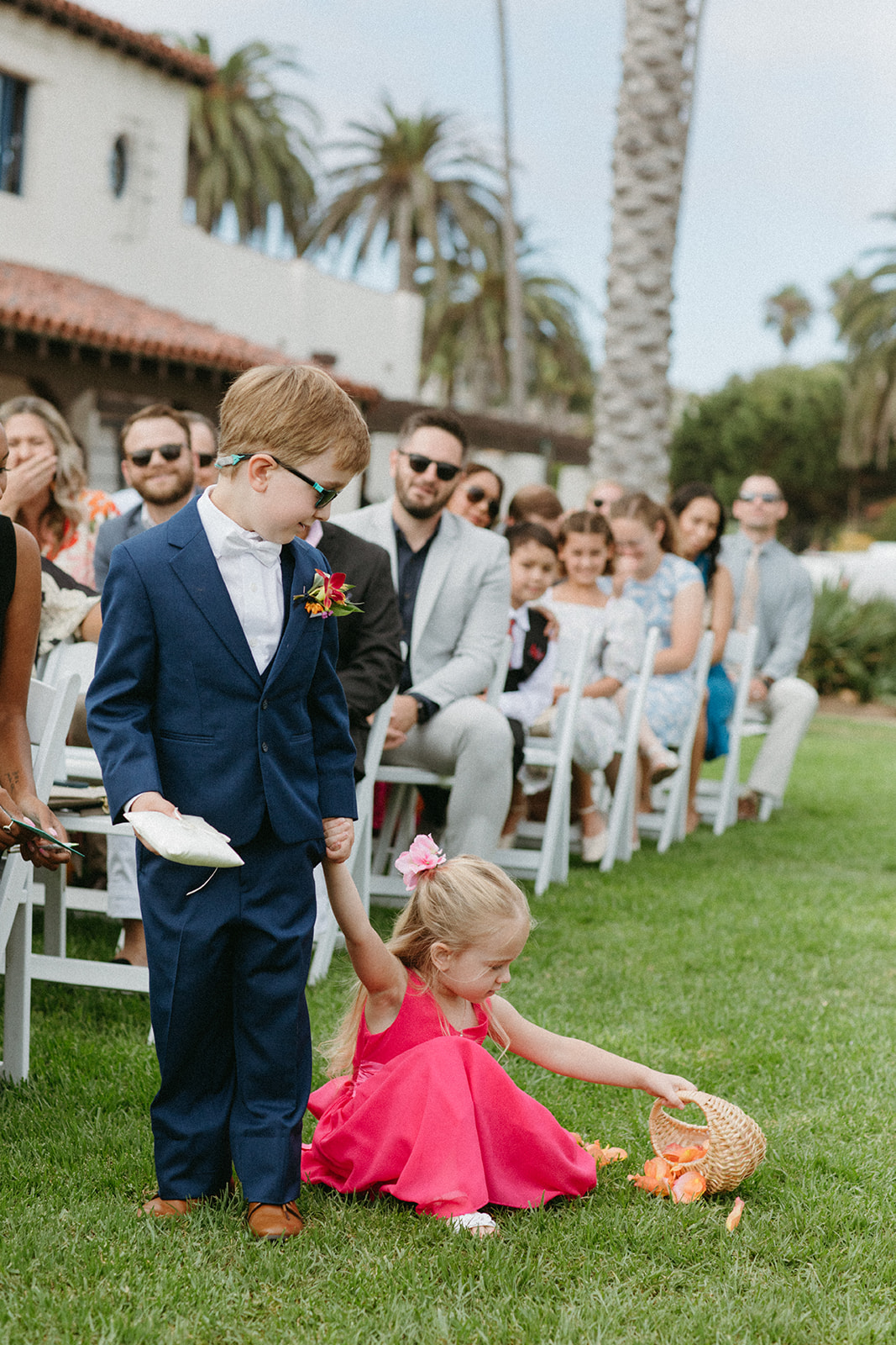 ring bearer in blue suit and white bow tie holds hands with flower girl in bright pink dress when she drops her basket on the ground 
