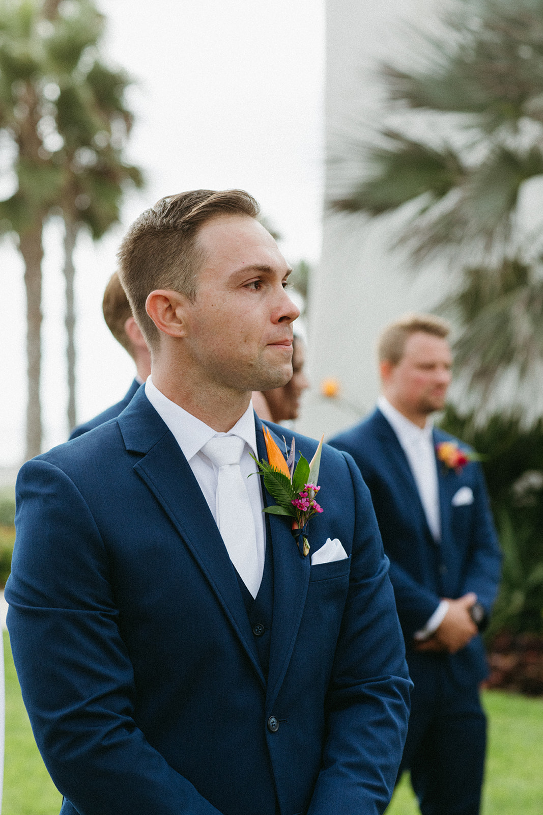 groom in blue suit and white tie and tropical boutonniere watches bride walk down aisle 