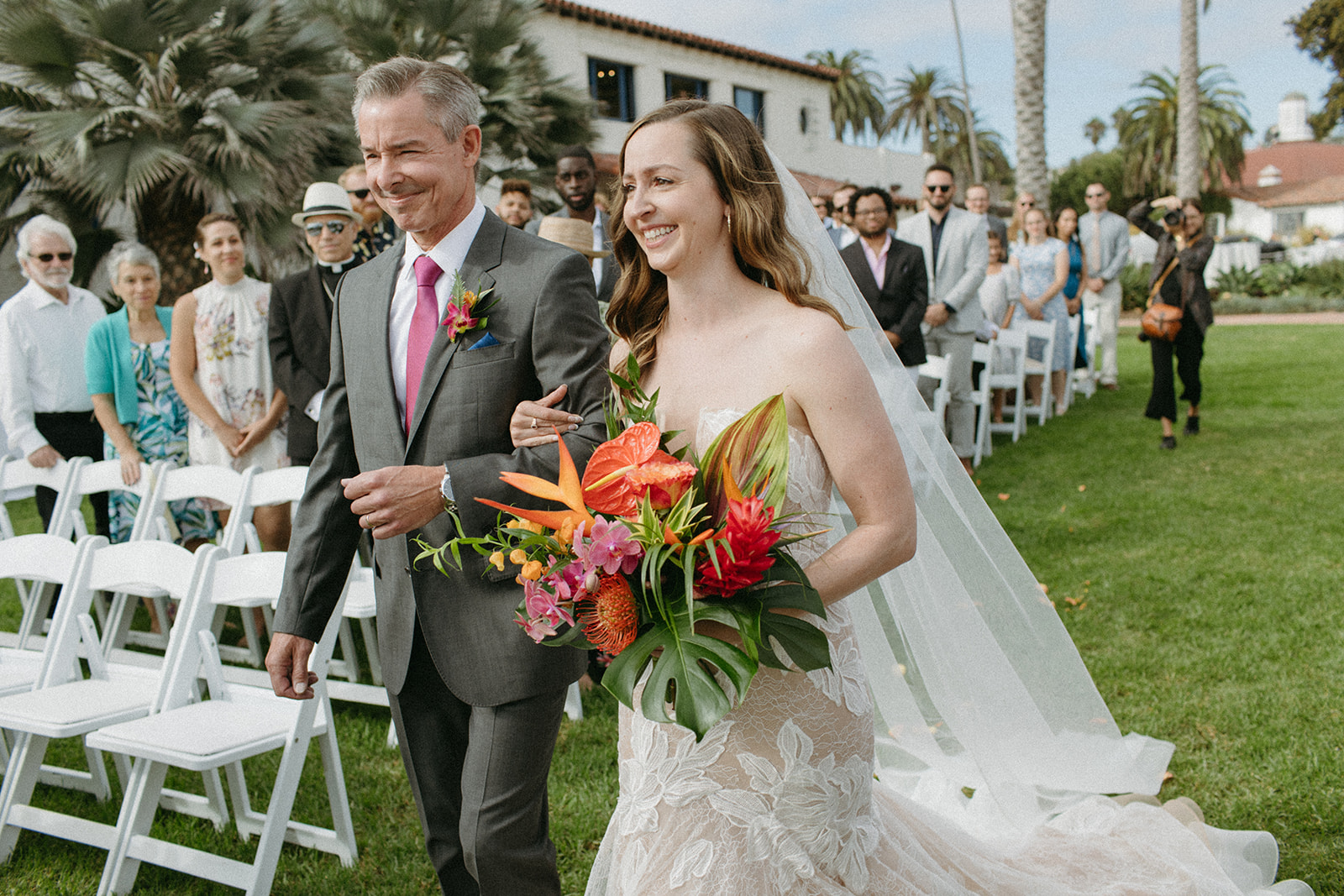 bride in strapless champagne dress with lace overlay walks down aisle with father of the bride in grey suit and bright pink tie