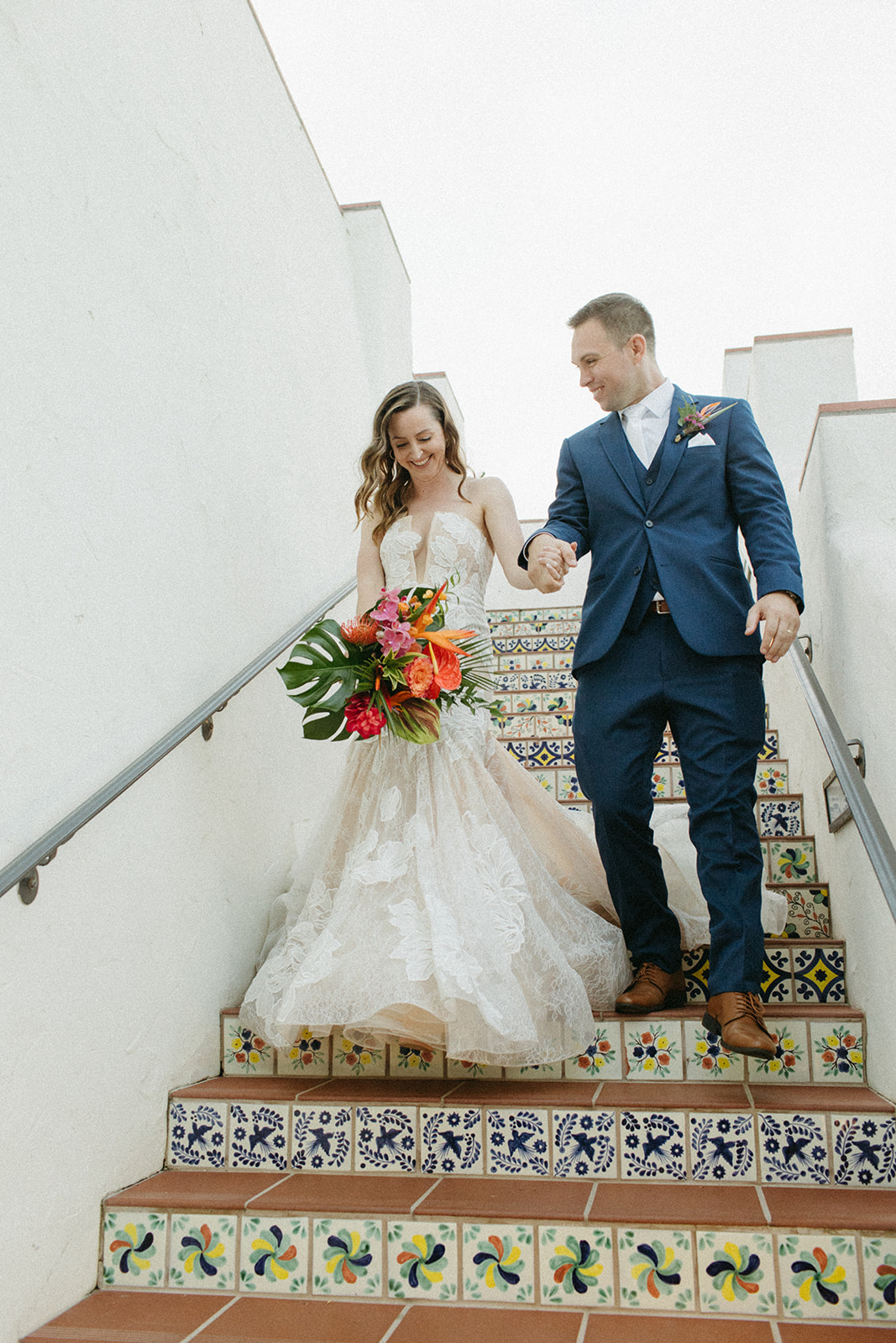bride in strapless champagne with lace overlay mermaid wedding dress and tropical bouquet walks with groom in blue suit with white tie down Spanish tiled staircase