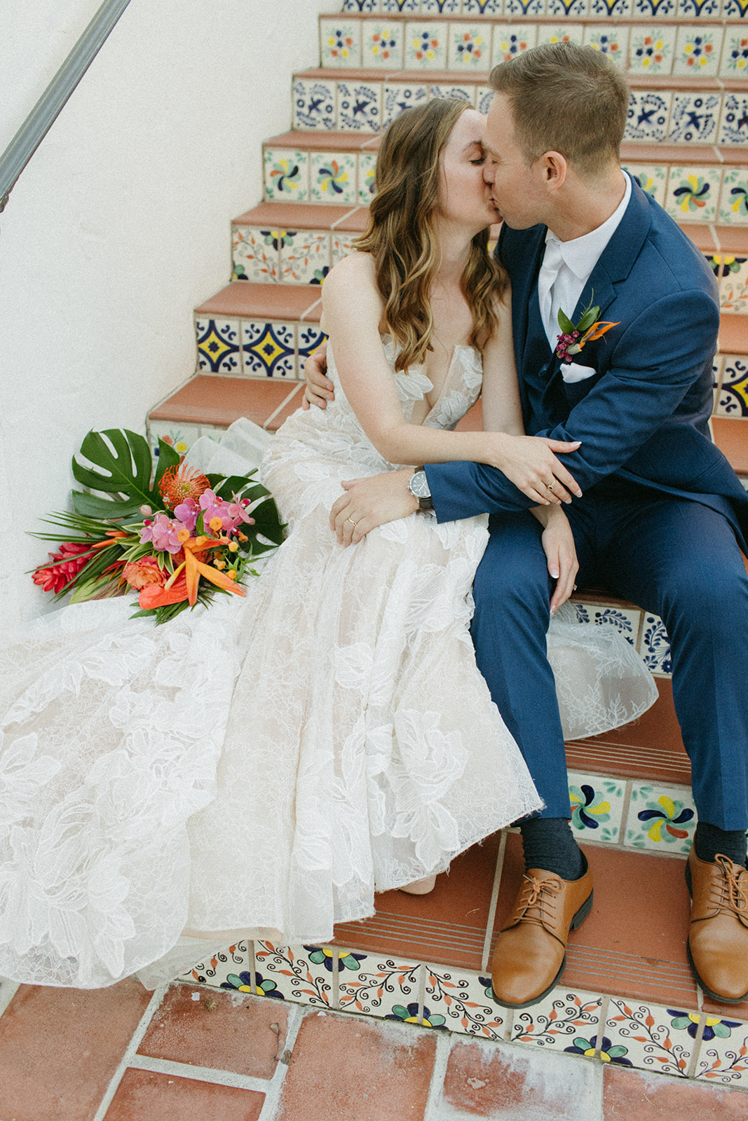 bride in strapless champagne with lace overlay mermaid wedding dress and tropical bouquet kisses groom in blue suit with white tie on Spanish tiled staircase