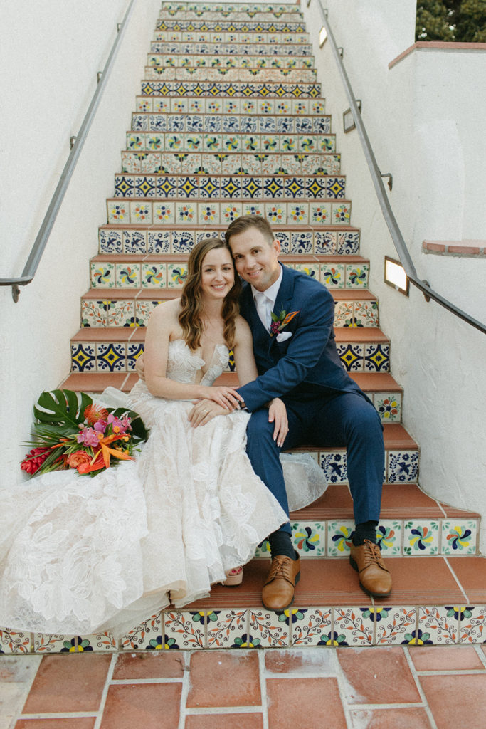 bride in strapless v neck lace dress and groom in blue suit sit on Spanish tile inspired staircase during their tropical themed wedding at Ole Hanson Beach Club