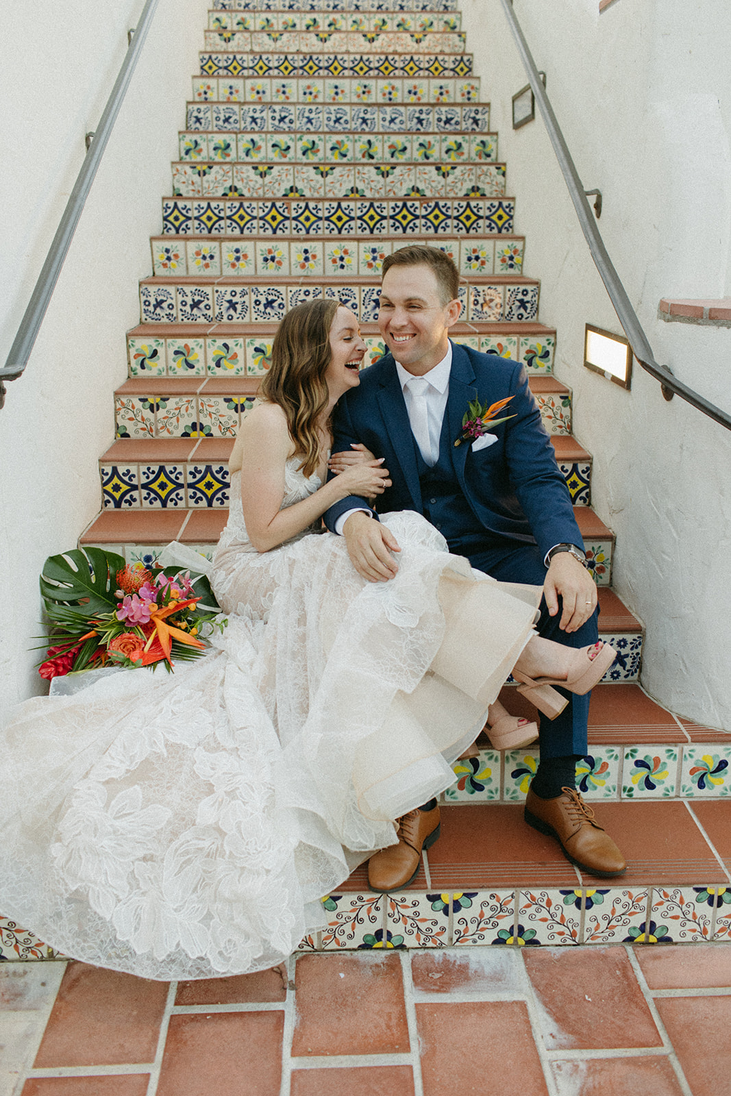bride in strapless champagne with lace overlay mermaid wedding dress and tropical bouquet with groom in blue suit with white tie on Spanish tiled staircase