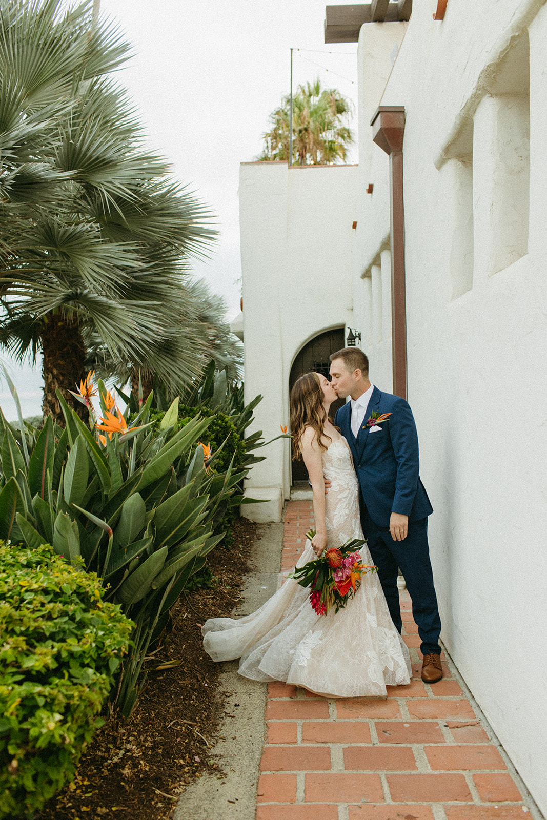 bride in strapless champagne with lace overlay mermaid wedding dress and tropical bouquet with groom in blue suit with white tie portrait shot overlooking the ocean