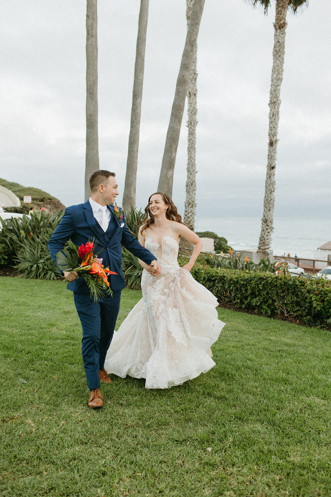 bride in strapless champagne with lace overlay mermaid wedding dress and tropical bouquet with groom in blue suit with white tie portrait shot overlooking the ocean