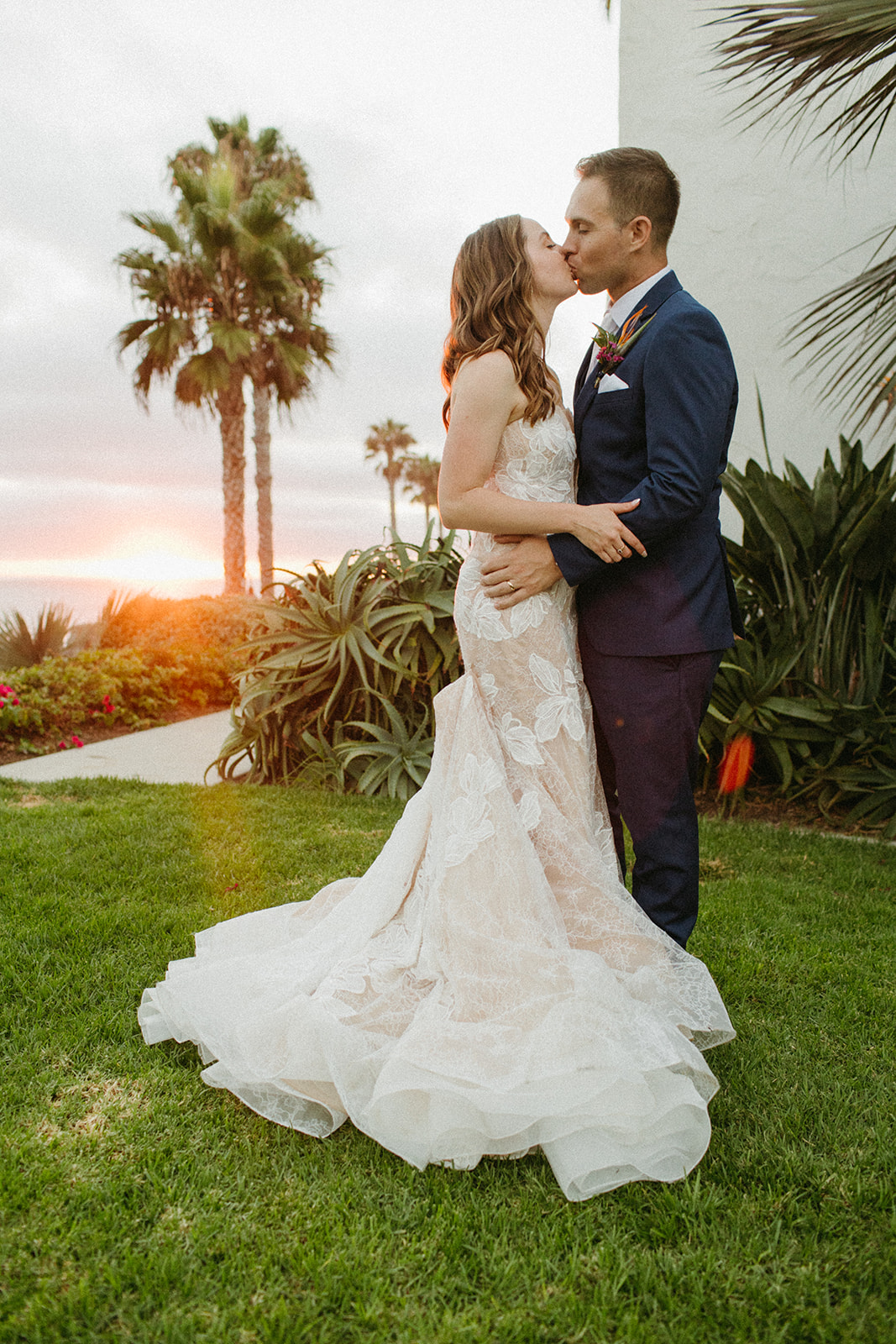 bride in strapless champagne with lace overlay mermaid wedding dress and tropical bouquet with groom in blue suit with white tie take sunset portrait shot overlooking the ocean