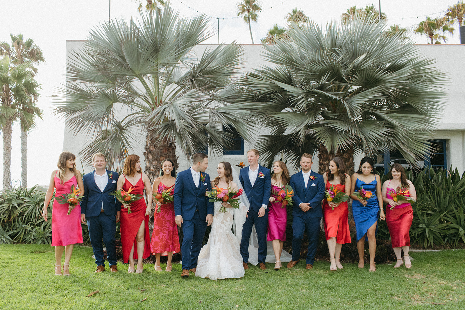 bride in strapless champagne with lace overlay mermaid wedding dress and tropical bouquet and groom in blue suit with white tie with wedding party in bright pink dresses and blue suits