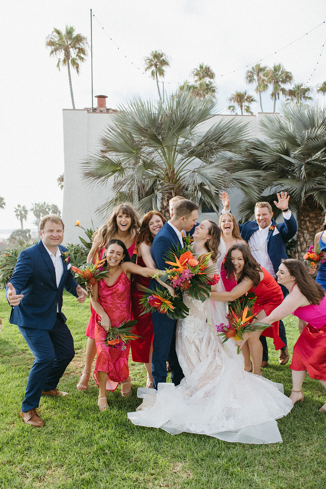 bride in strapless champagne with lace overlay mermaid wedding dress and tropical bouquet and groom in blue suit with white tie with wedding party in bright pink dresses and blue suits