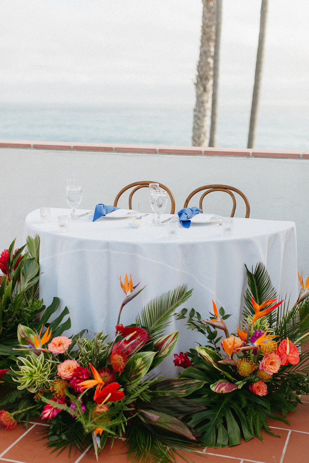 sweetheart table with wooden chairs and white linens, blue napkins and tropical floral table arrangements