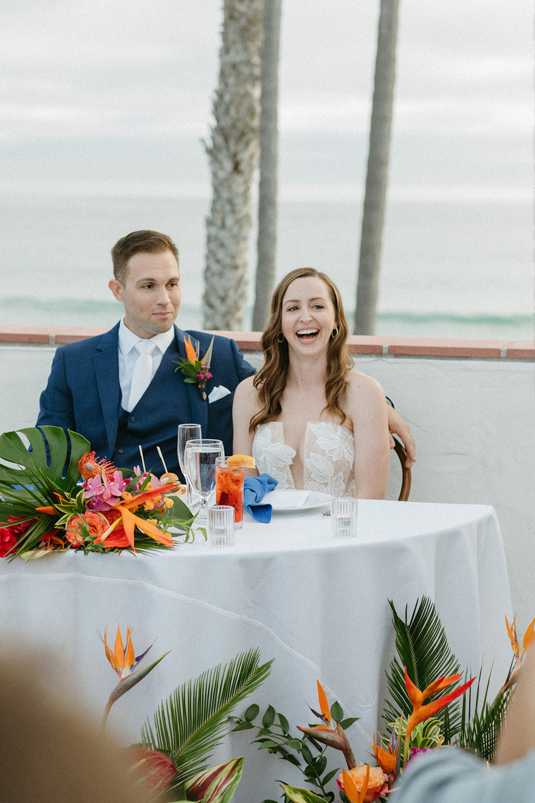 bride and groom sit at sweetheart table during outdoor wedding reception with ocean in the backdrop