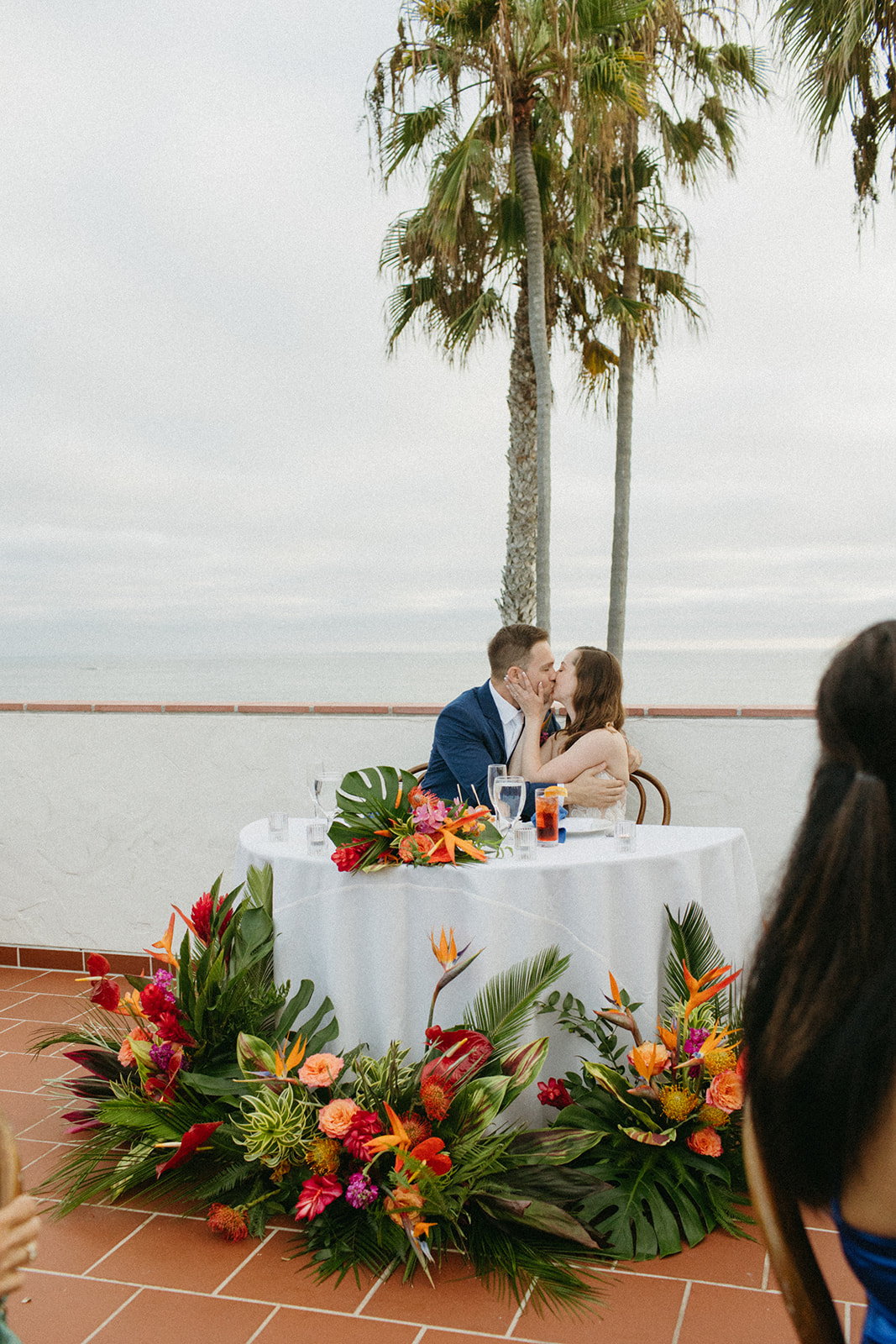 bride and groom kiss at sweetheart table with tropical floral arrangements and ocean in the background