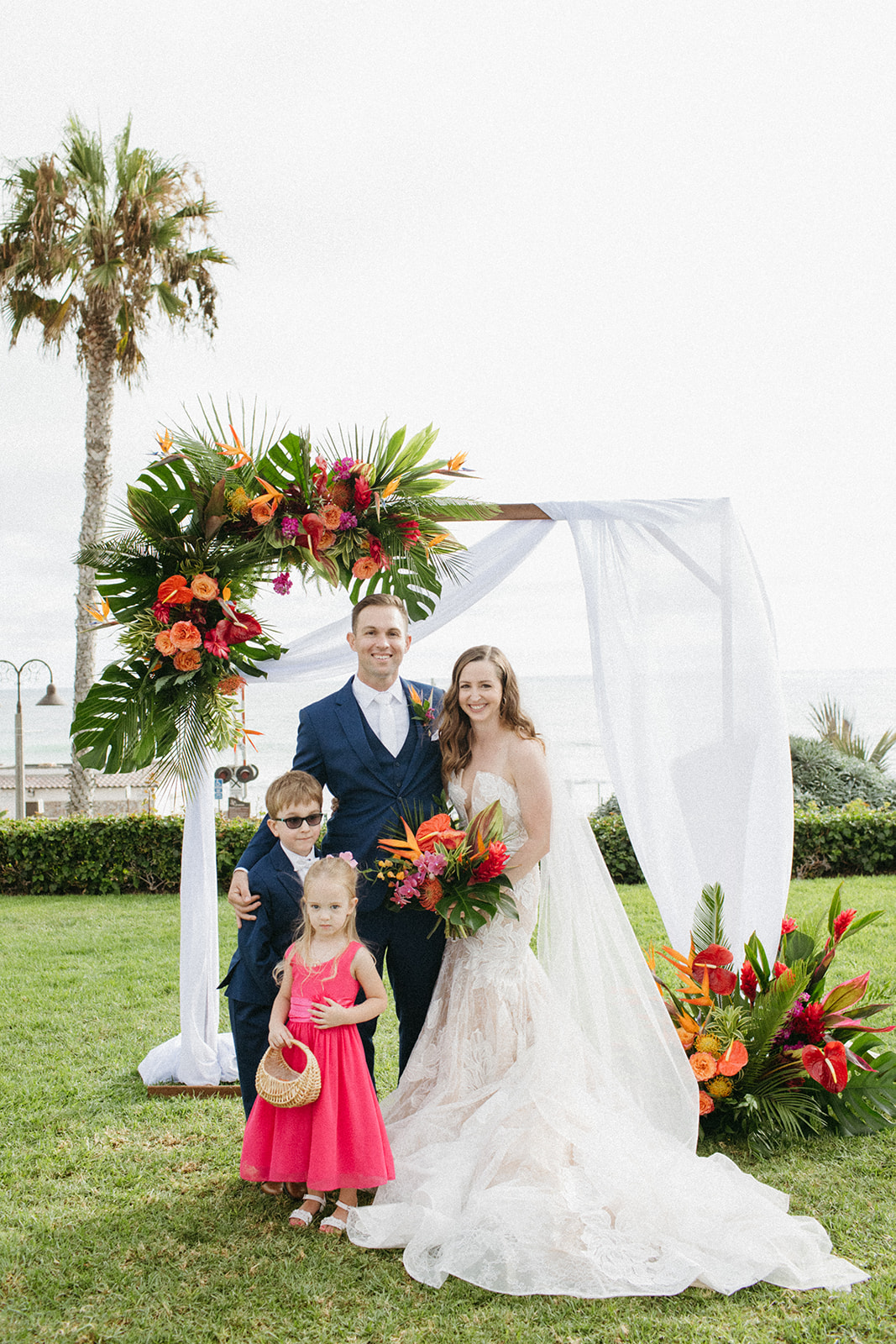 bride and groom with ring bearer and flower girl stand in front of their tropical inspired ceremony arch