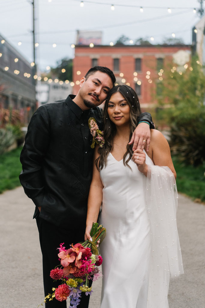 bride in modern silk white wedding dress with pearls in her hair holding bright bridal bouquet stands with groom in all black suit outside The Revery with market lights behind them