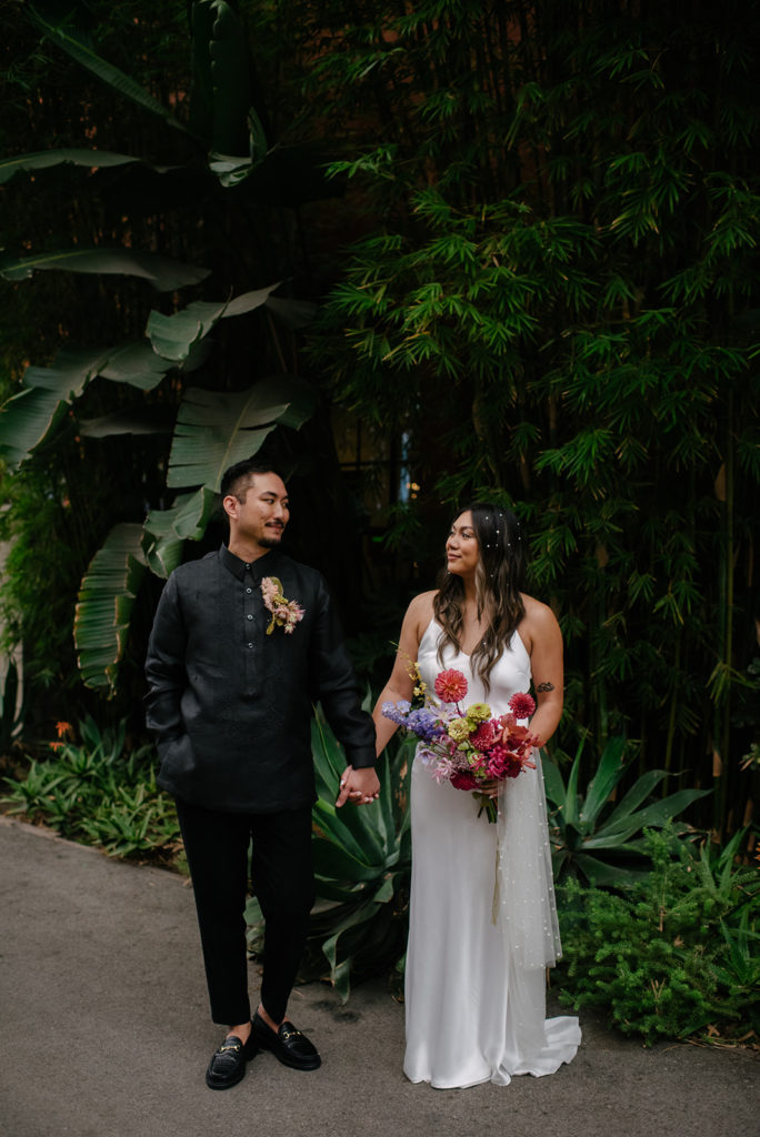 bride in modern silk dress with pearls in hair and colorful bouquet stands with groom in all black suit and bold boutonniere surrounded by greenery at The Revery