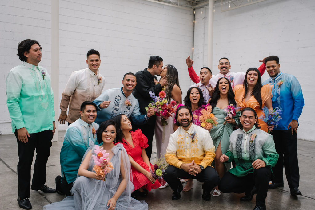 bride in modern silk wedding dress and groom in all black suit stands with co-ed wedding party in colorful rainbow inspired outfits